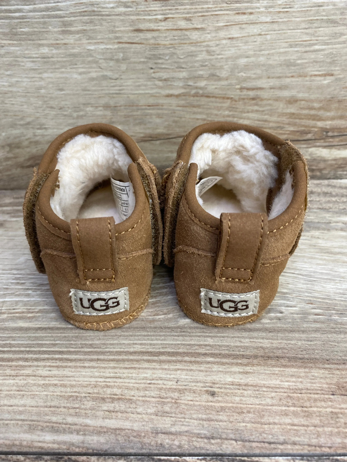 Brown UGG baby booties with white fleece lining on wooden surface, cozy kids footwear