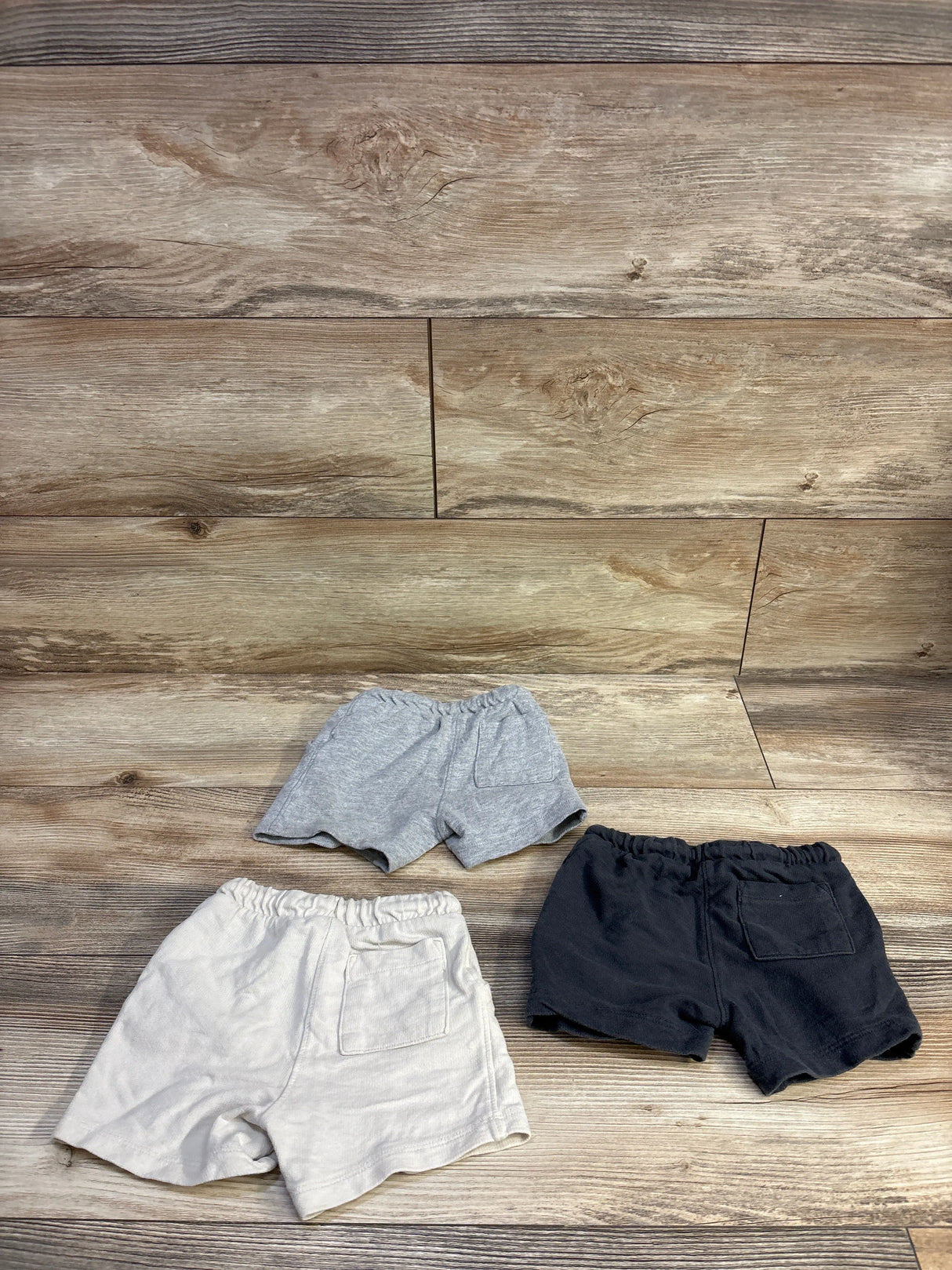 Top-down view of three toddler shorts (light gray, cream, black) showing elastic waists and back patch pockets on wooden floor