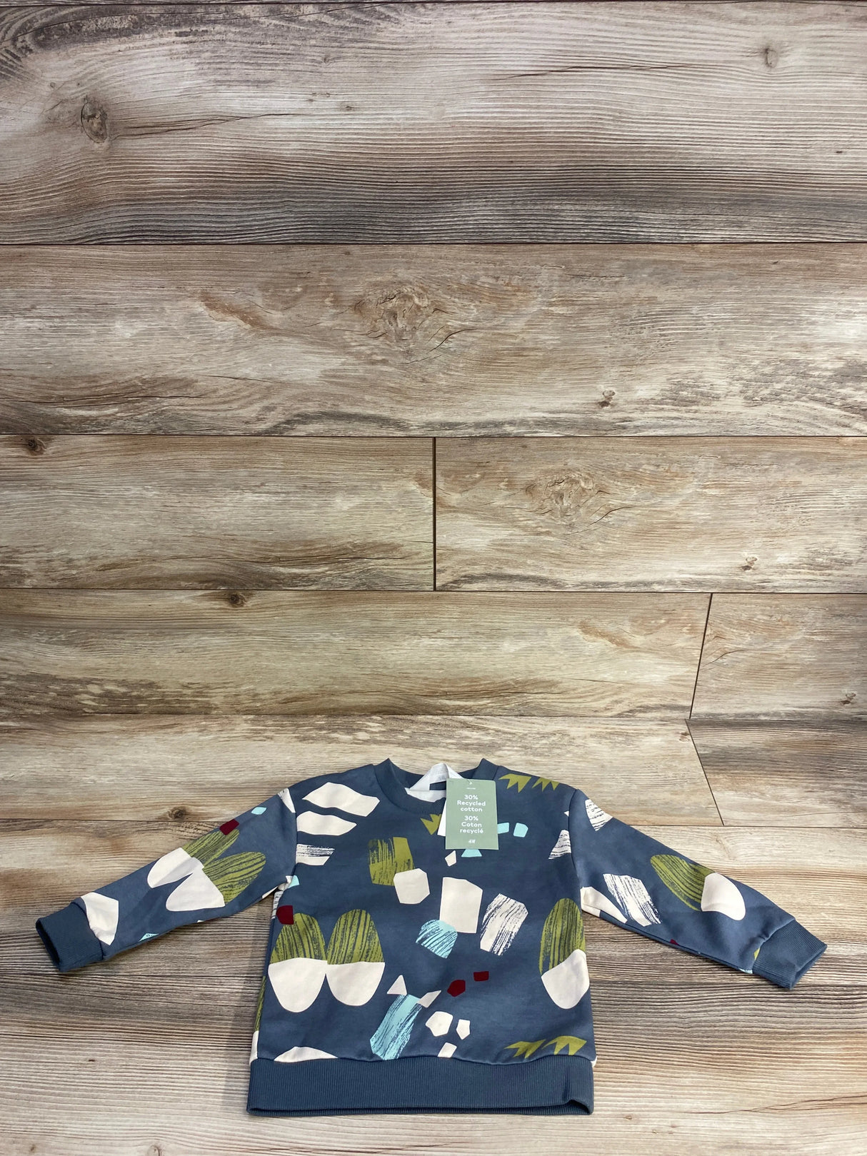 Flat lay of H and M light blue toddler sweatshirt (2T) showing abstract green, white, and yellow print and ribbed cuffs on wood background