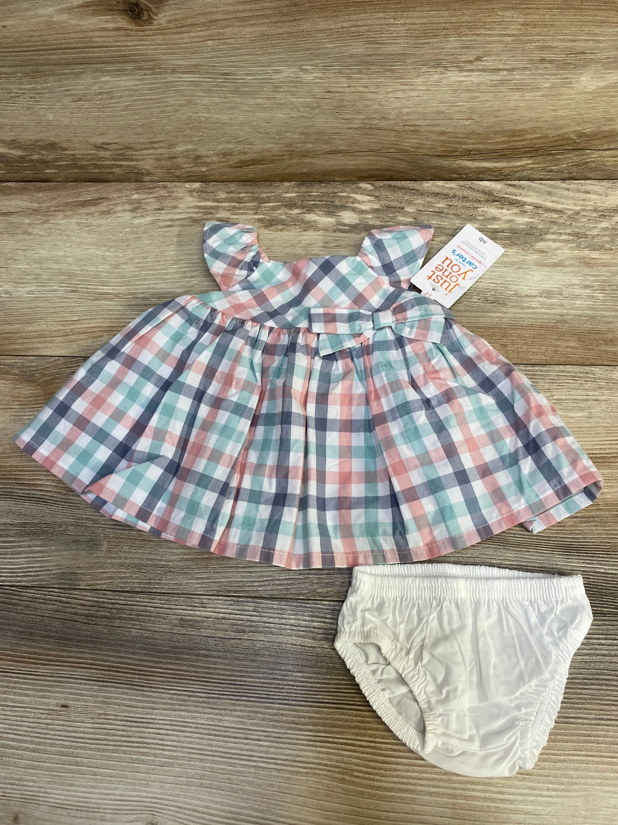 Top-down view of newborn plaid dress and white bloomers showing ruffled cap sleeves, waist bow, and pastel pink-blue-green check on wood surface