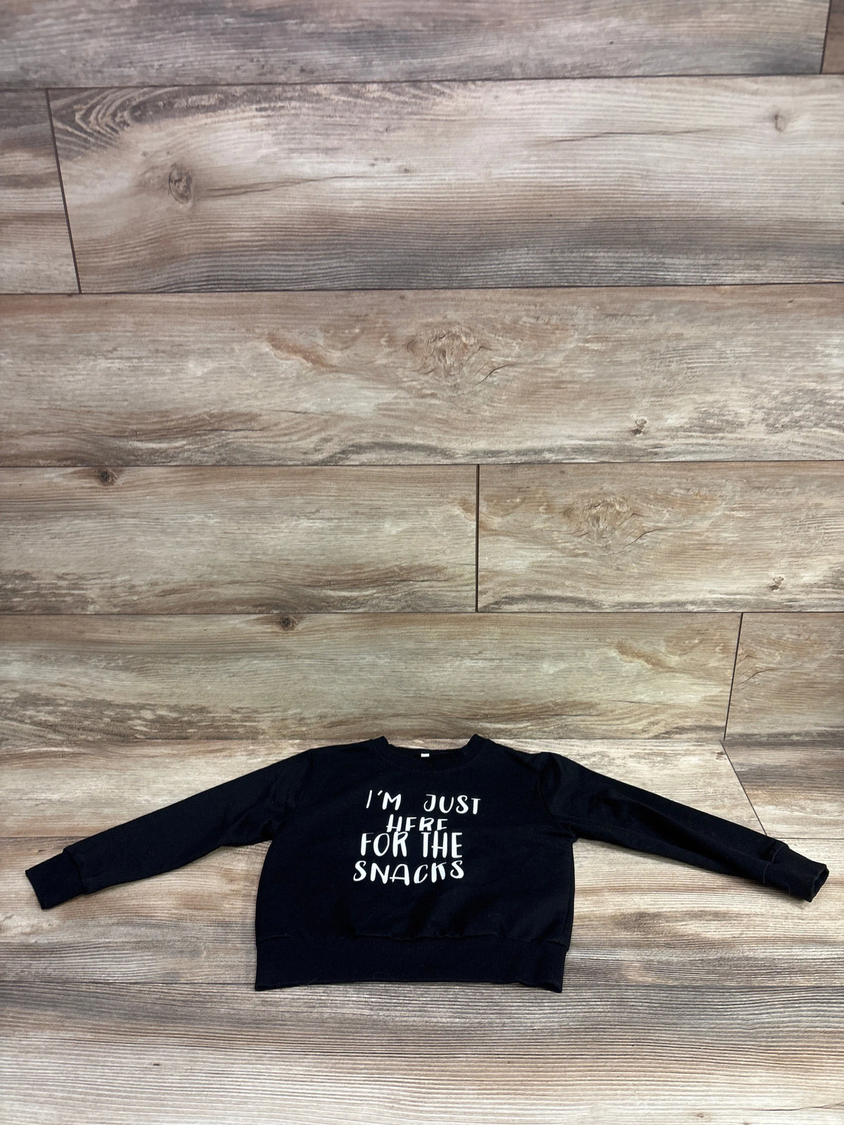Front view of black pullover sweatshirt for ages 18 to 24 months with white I am just here for the snacks text on wood floor