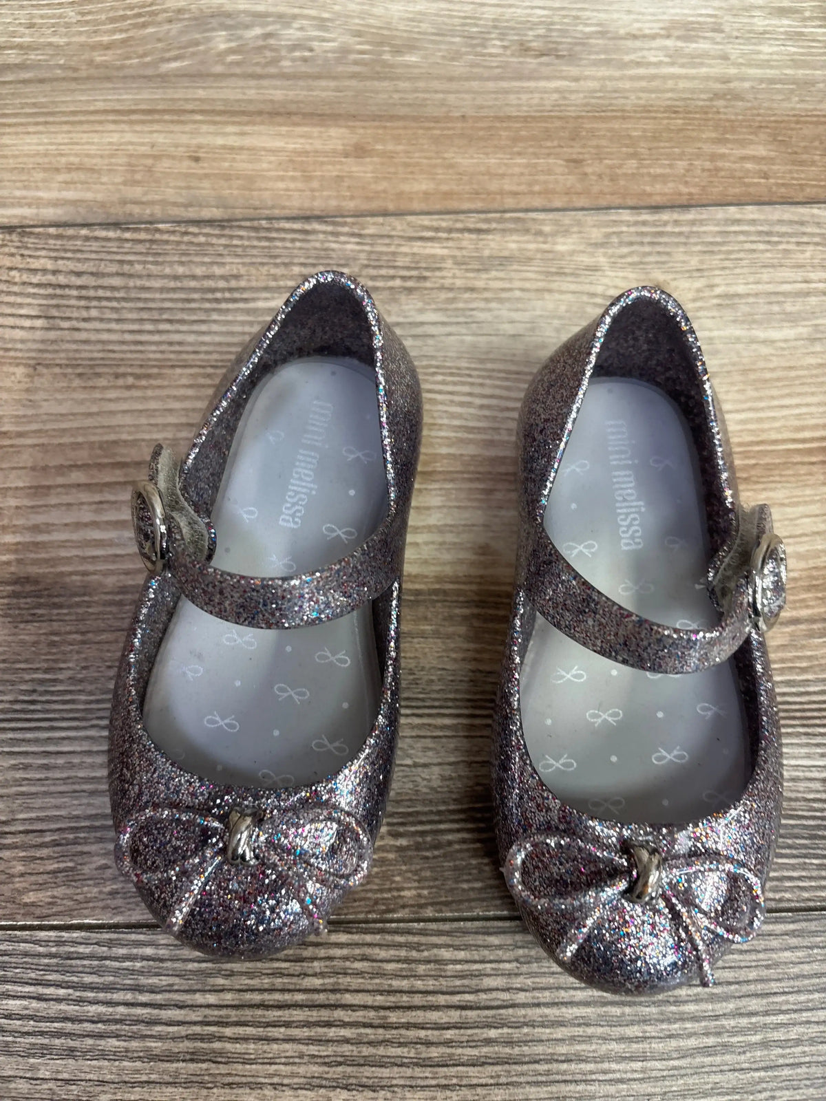 Top view of Mini Melissa Sweet Love jelly flats size 7C showcasing silver multicolor glitter finish, bow detail, and Mary Jane strap with wood floor background