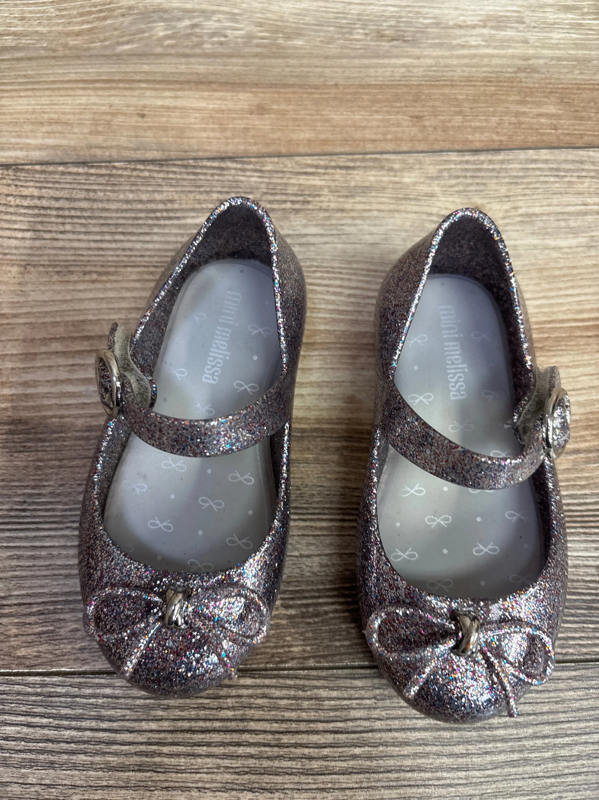 Top view of Mini Melissa Sweet Love jelly flats size 7C showcasing silver multicolor glitter finish, bow detail, and Mary Jane strap with wood floor background
