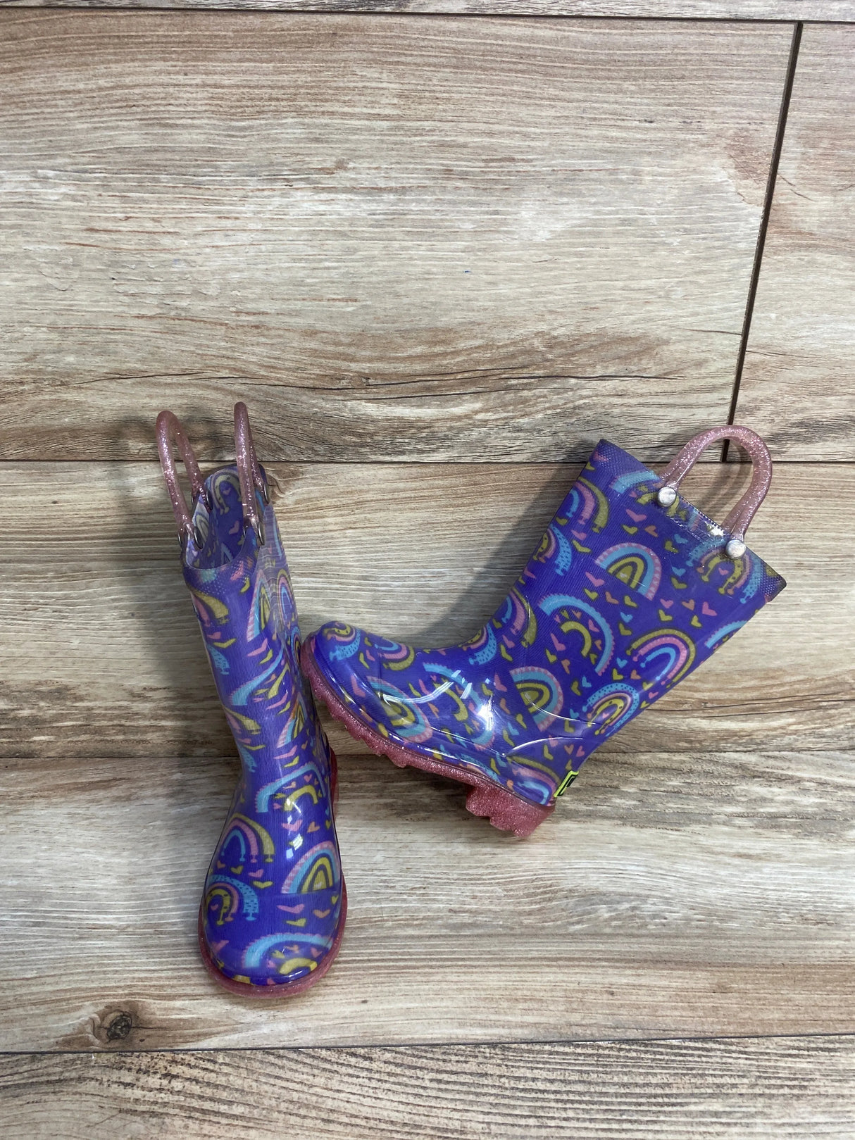 Side view of Western Chief Kids light-up rain boots in purple with rainbow print and pink glitter pull handles on wood plank floor