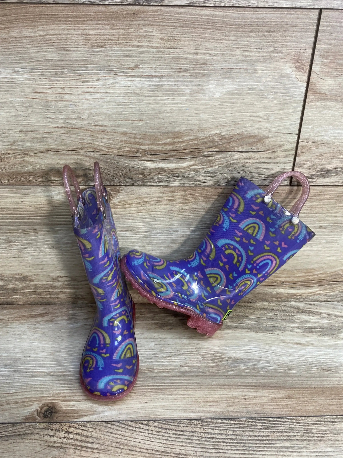Front view of Western Chief kids light up rain boots showing purple rainbow-and-heart pattern and pink glitter pull handles on wood floor