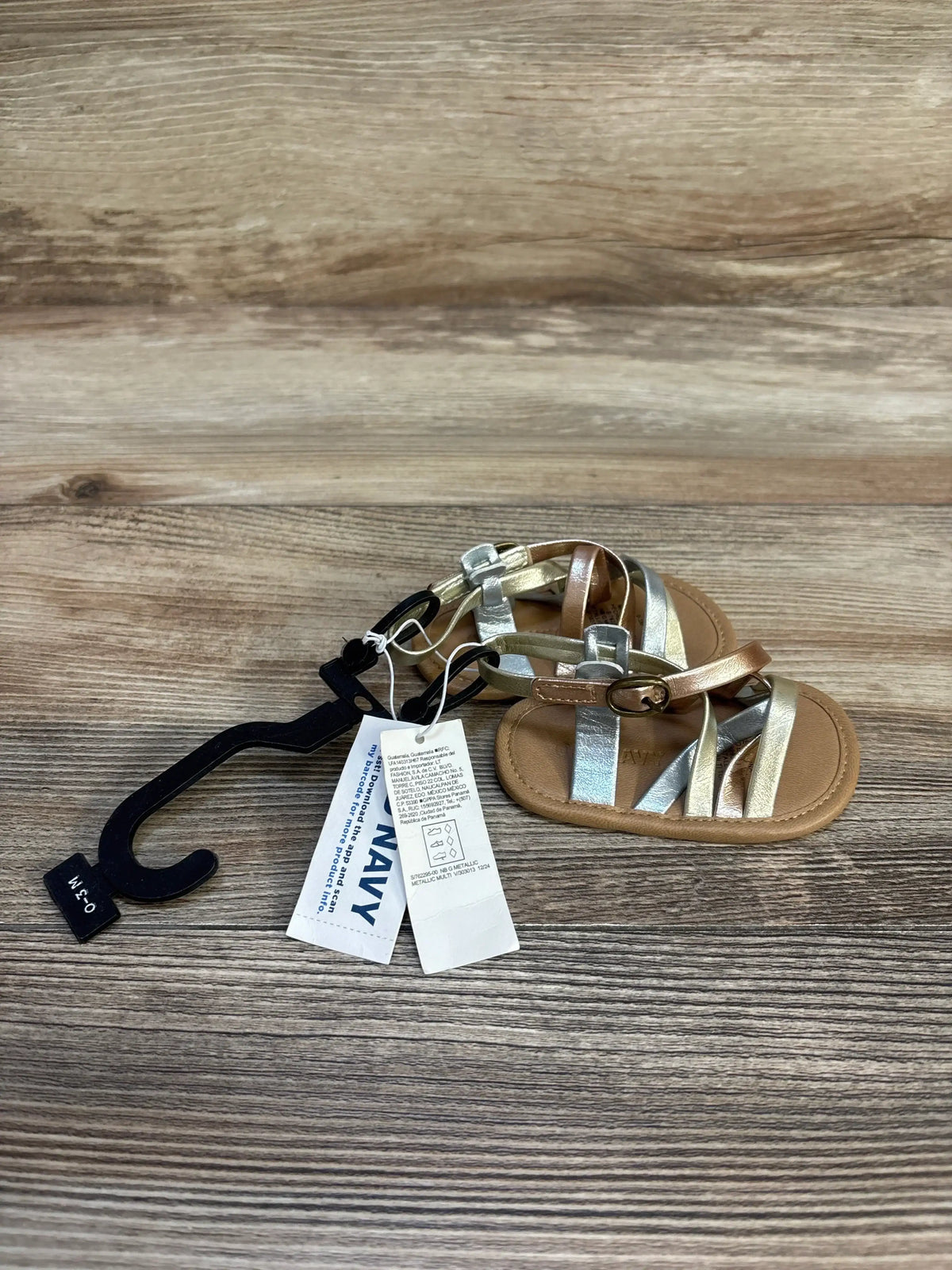 Metallic baby sandals with gold and silver straps, tags attached, on wooden surface