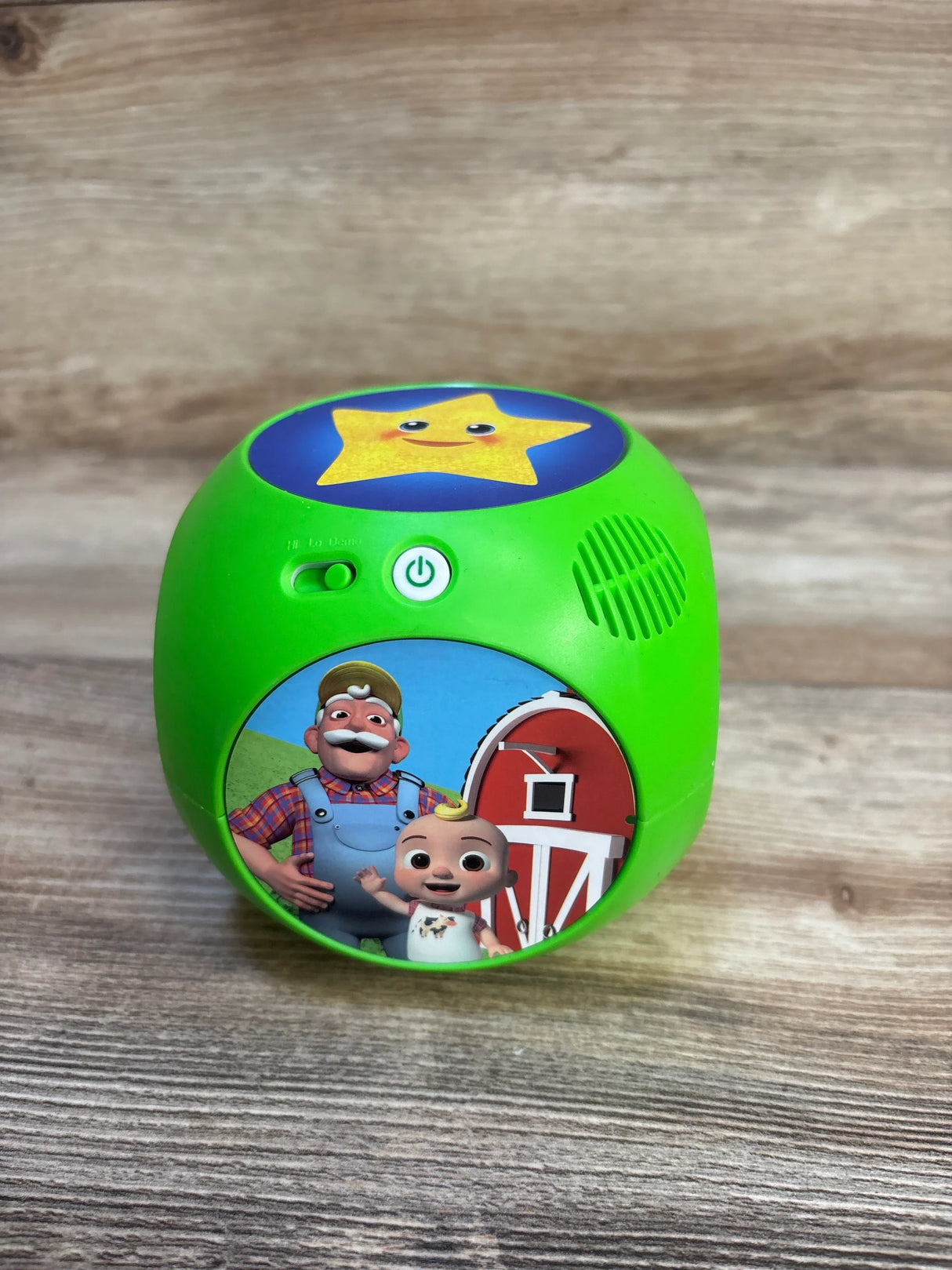 Front of Cocomelon Tumble Tunes toy, green cube with power button, speaker grill and farm graphic on wood background