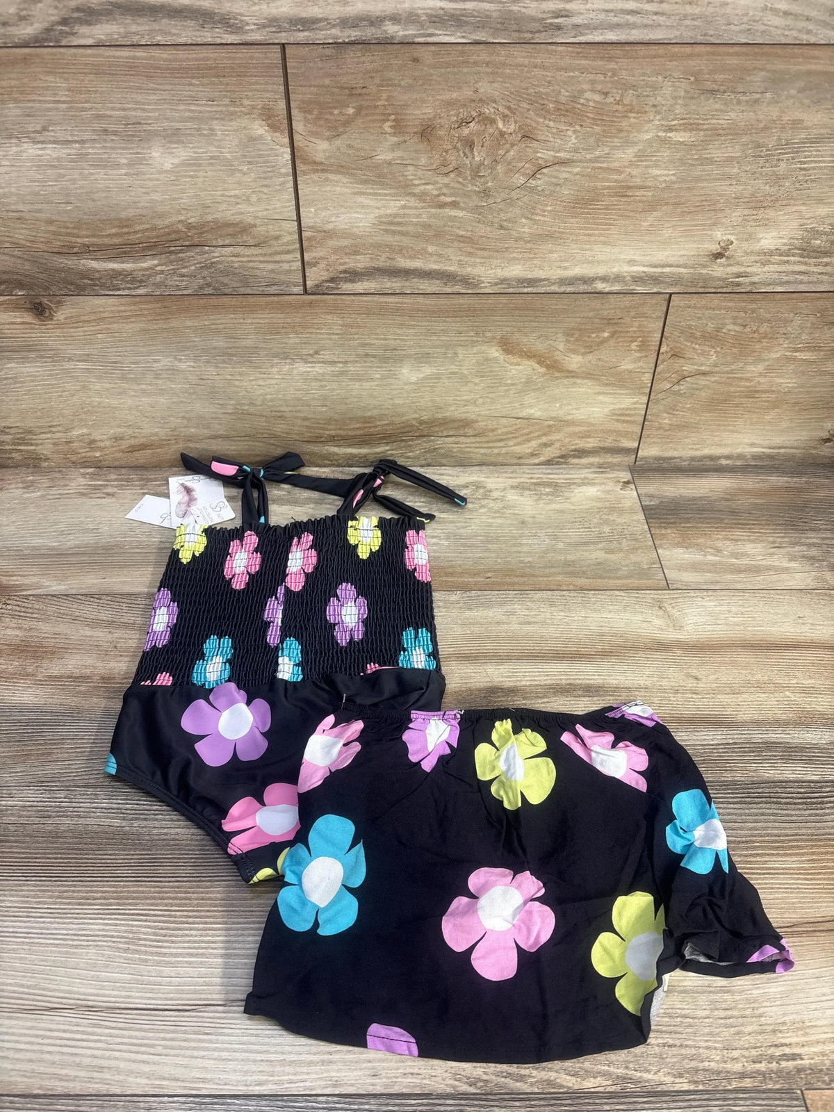 Top-down view of Jessica Simpson smocked swimsuit and skirt set with colorful floral print on black fabric and wood floor