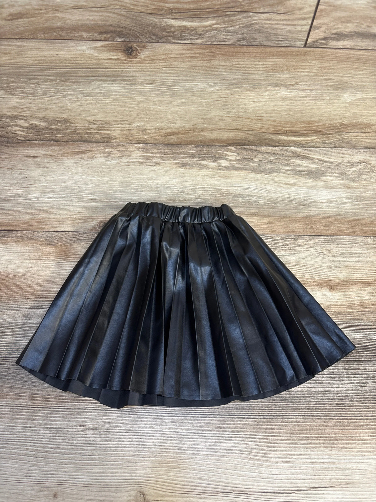 Black pleated faux leather skirt for kids, laid flat on wood floor