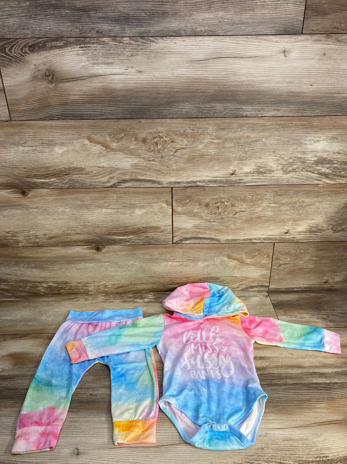 Top-down view of pink tie-dye hooded bodysuit and matching leggings showing the Little Miss Sassy Pants chest print on wood floor