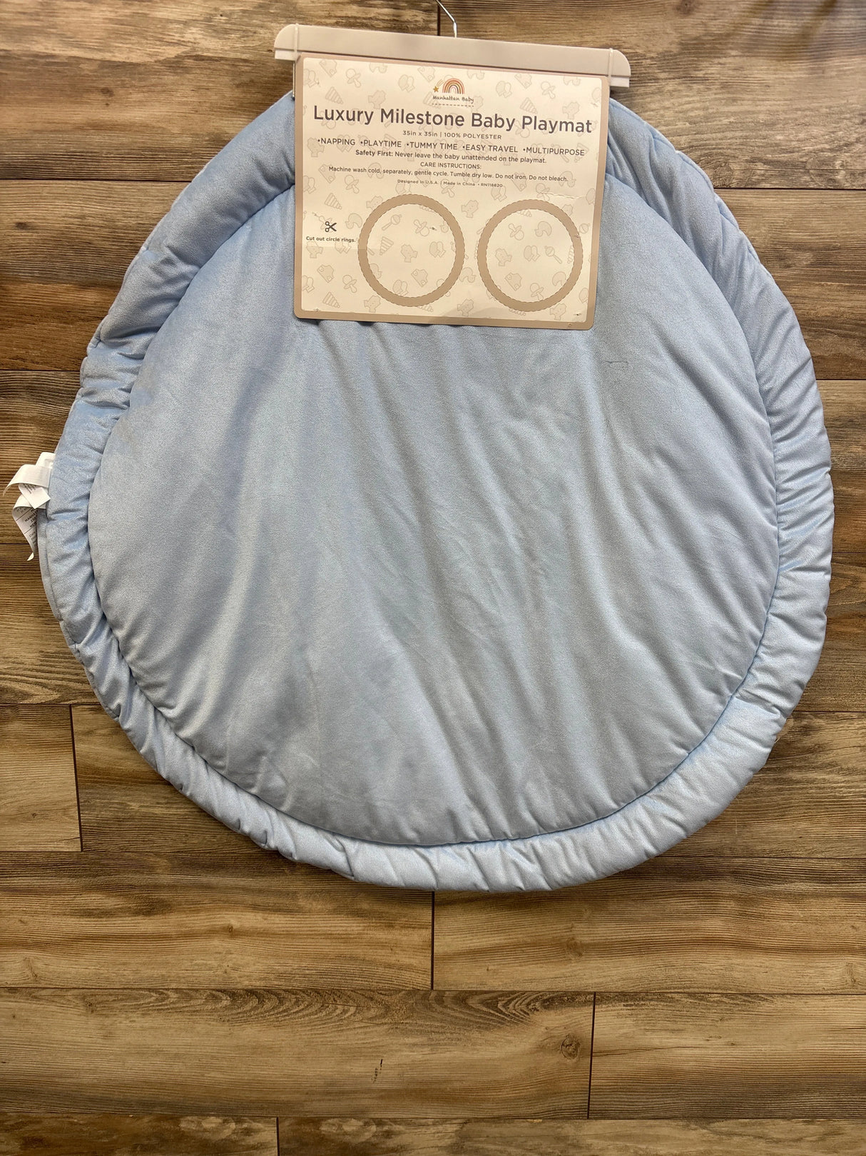 Top-down view of round light blue milestone baby playmat showing padded ruched edge and attached label on wood floor