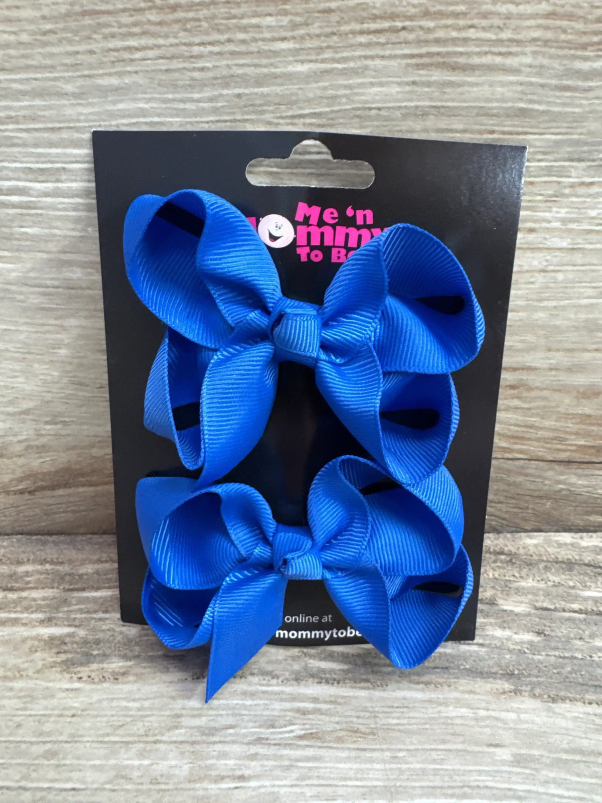 Front view of two medium blue grosgrain hair bows on a black branded display card against a light wood background