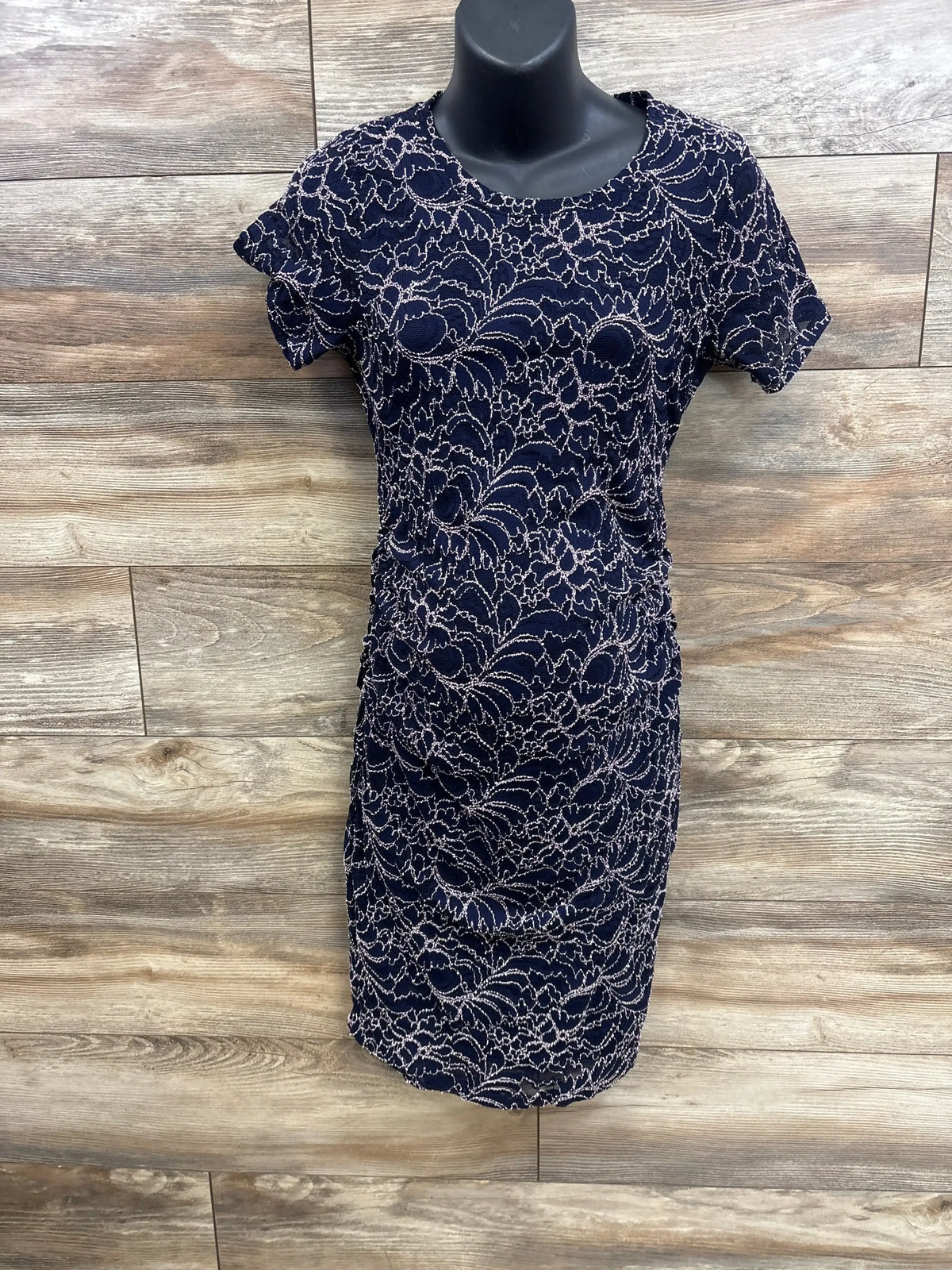 Front view of navy maternity lace dress showcasing short sleeves, fitted ruched sides, and knee-length hem on a black mannequin against wood plank background