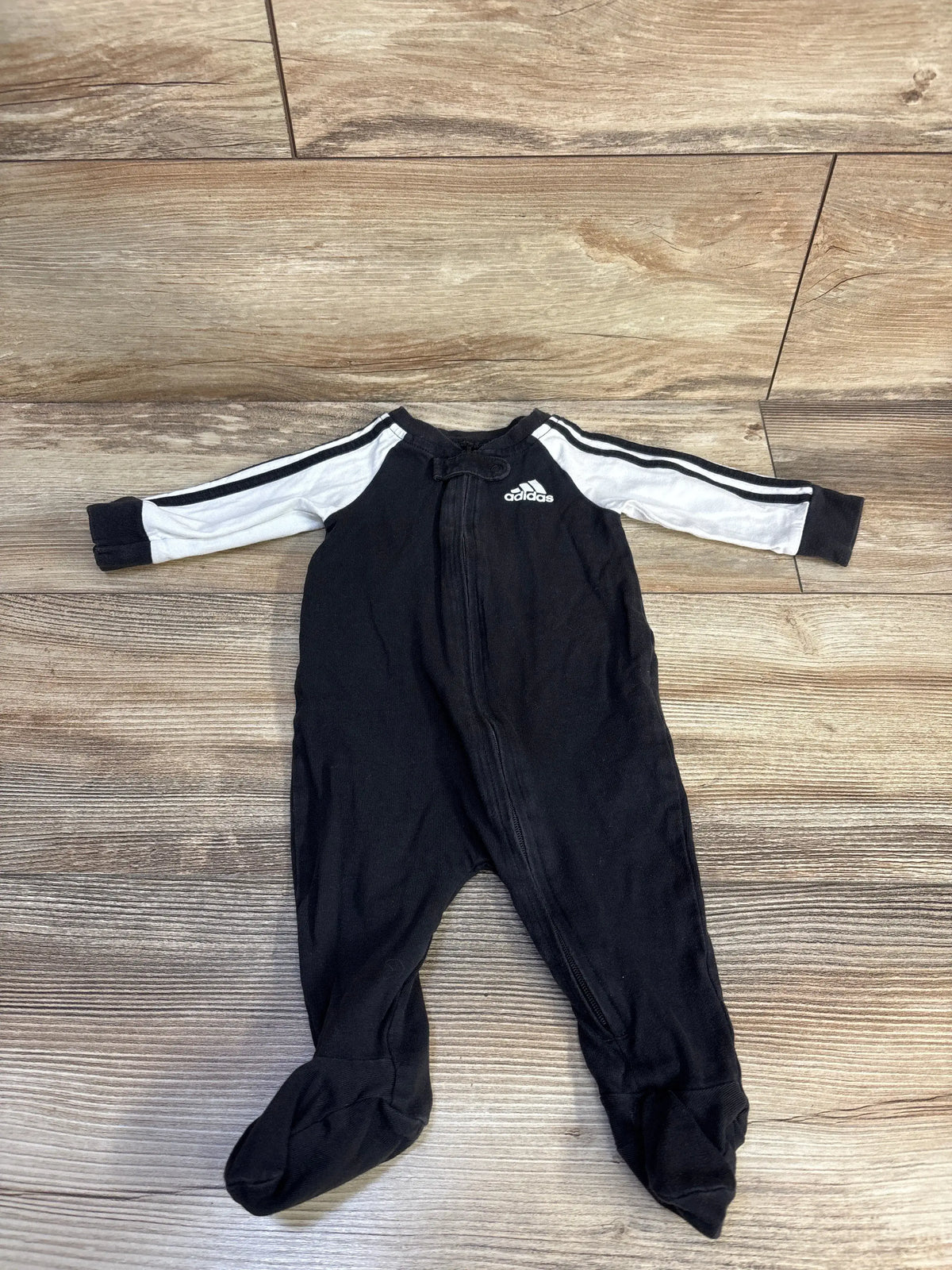 Front view of Adidas black 3-month baby sleeper with white striped sleeves, front zipper, logo, and footed legs on wood-look floor