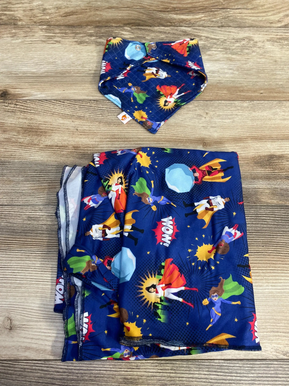 Top view of Bumblito stretch swaddle set Never Alone showing folded superhero-print swaddle and matching snap bib on wood