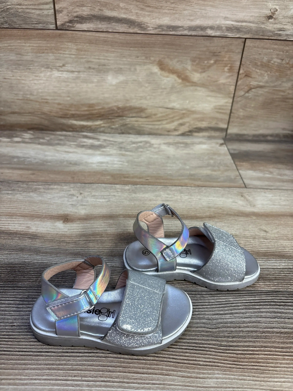 Front and rear views of Kensie Girl glitter sandals in silver showcasing ankle straps and hook and loop closures with a wood-look floor background