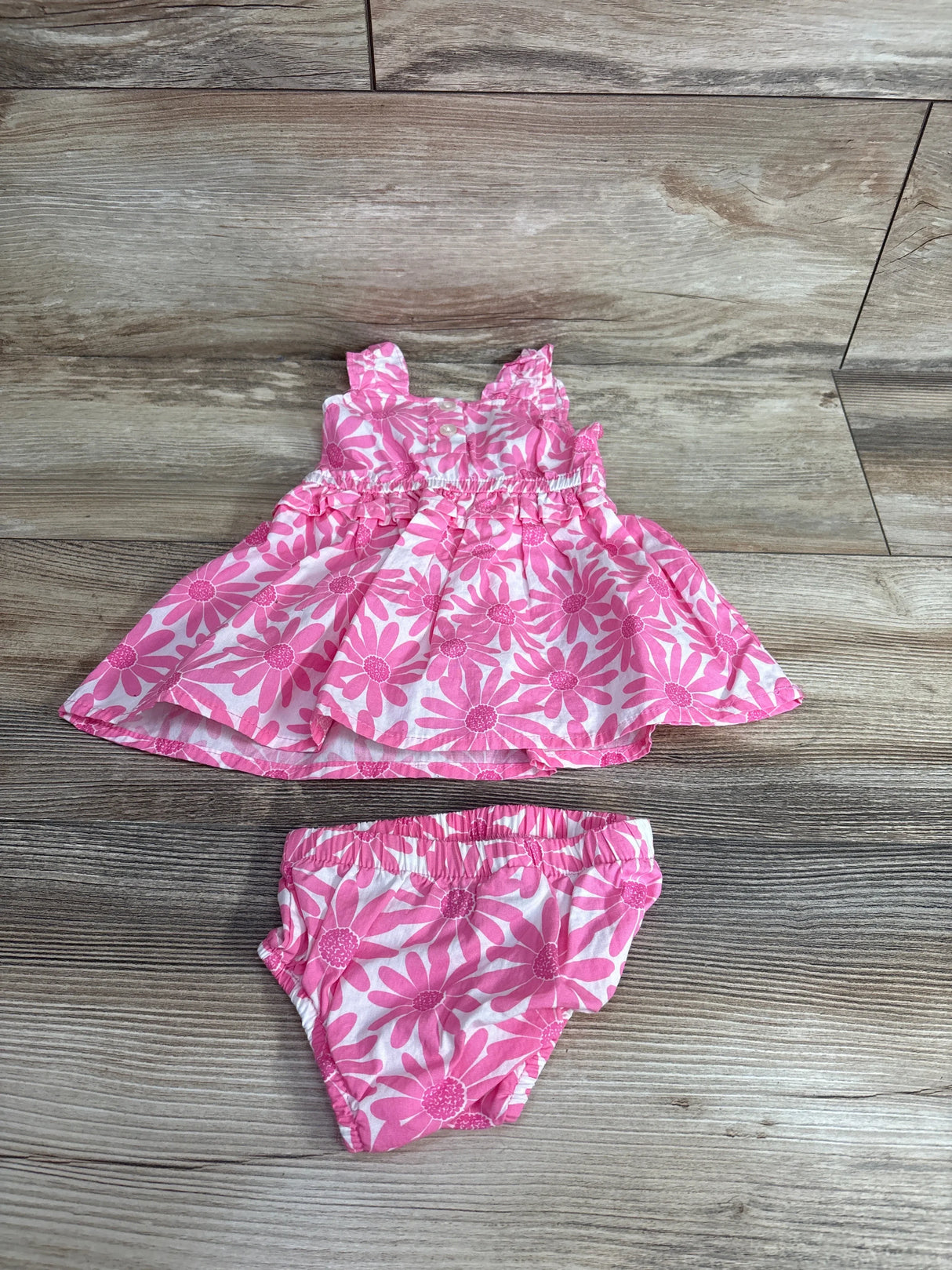 Top down view of pink floral baby dress and matching bloomers showing daisy print, ruffled straps and elasticized waist on wood floor