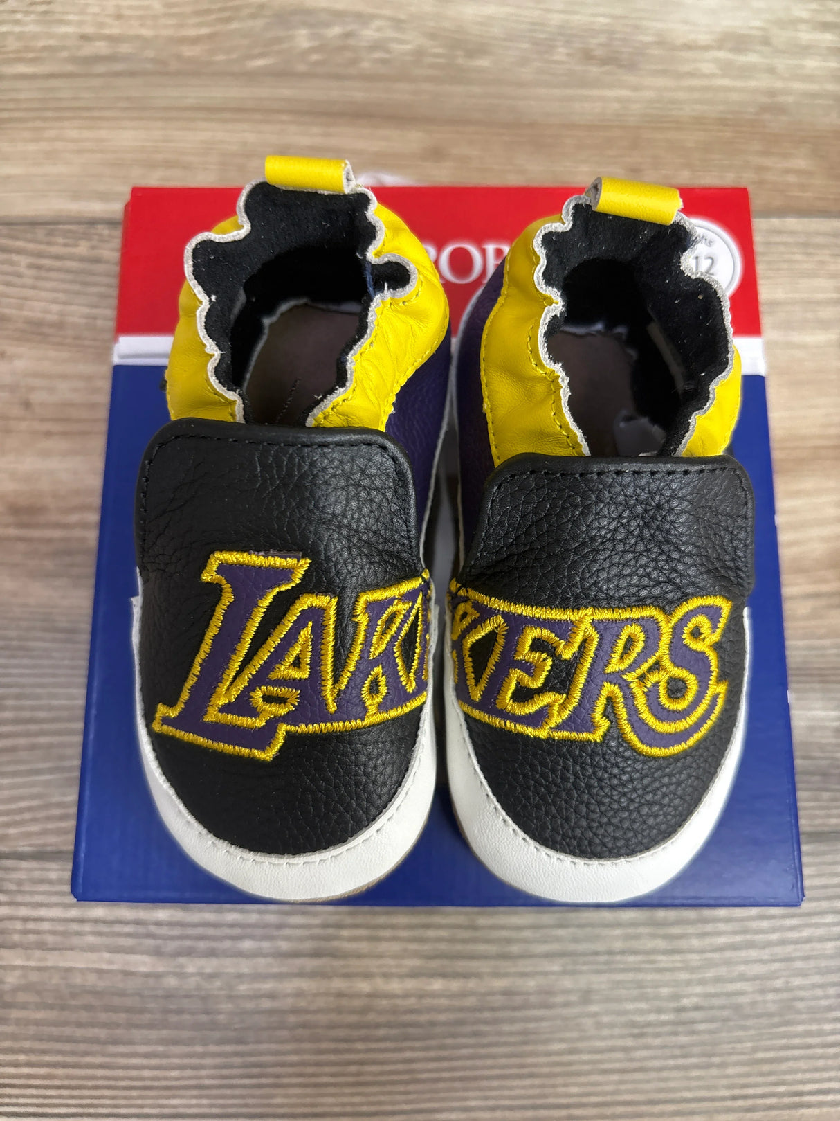 Baby shoes with black, yellow, and white colors, feature embroidered LA Lakers logo, on a box