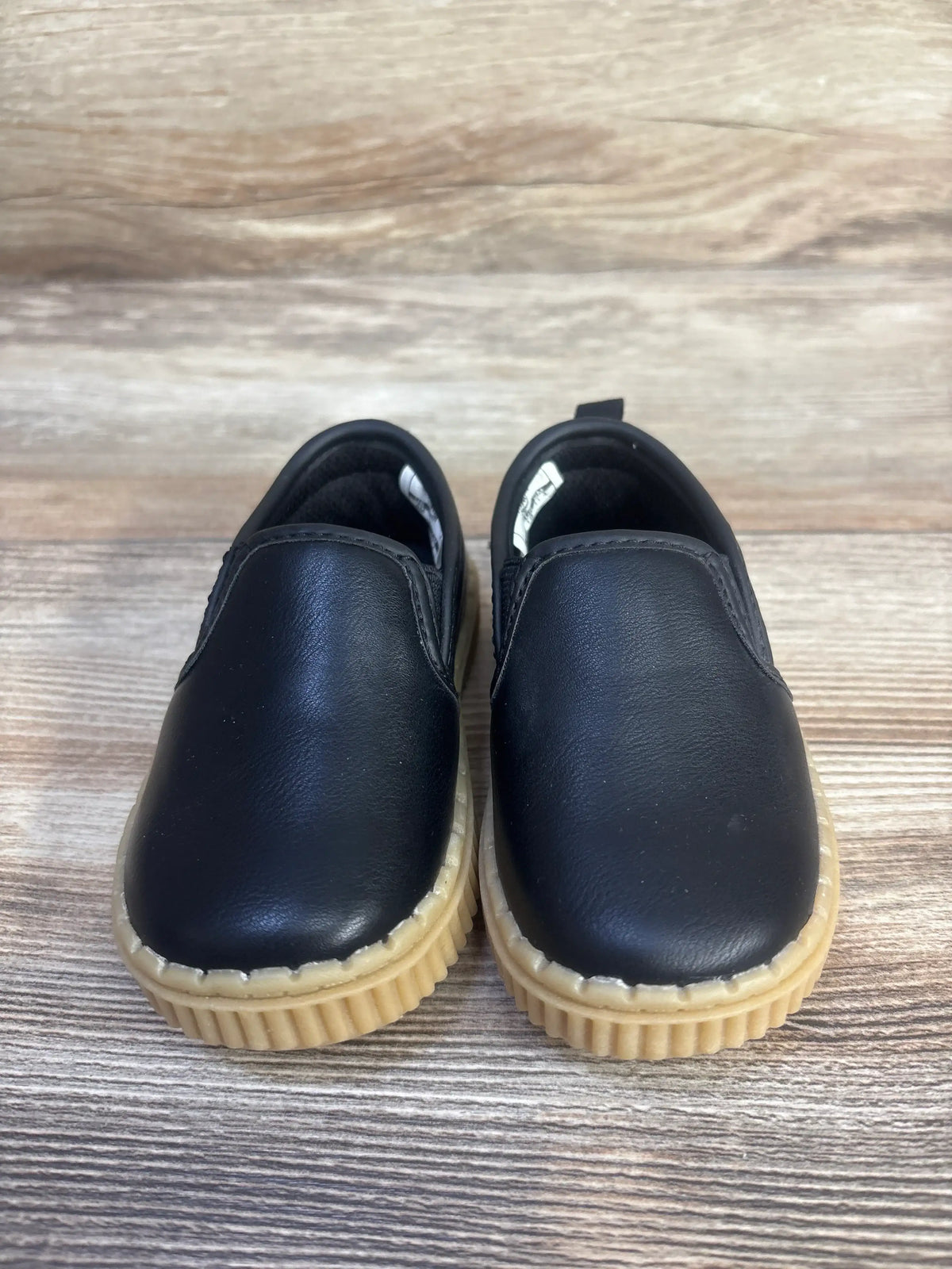 Front view of OshKosh Victor slip-on sneakers in black showcasing tan ribbed soles on a wood-grain background
