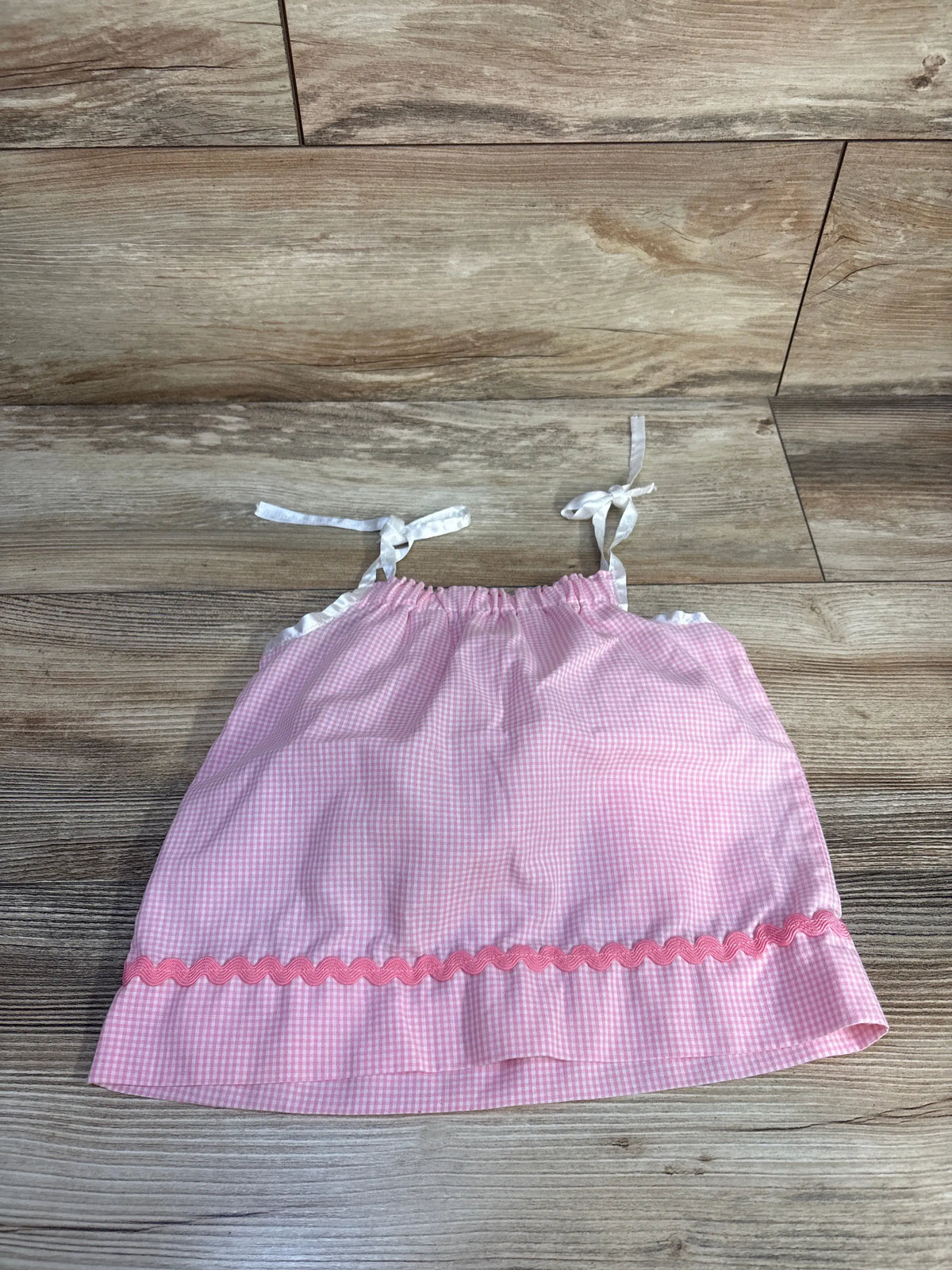 Front view of pink gingham sleeveless sundress in size 6 to 9 months showcasing tie shoulder straps and ruffled hem on a wood floor