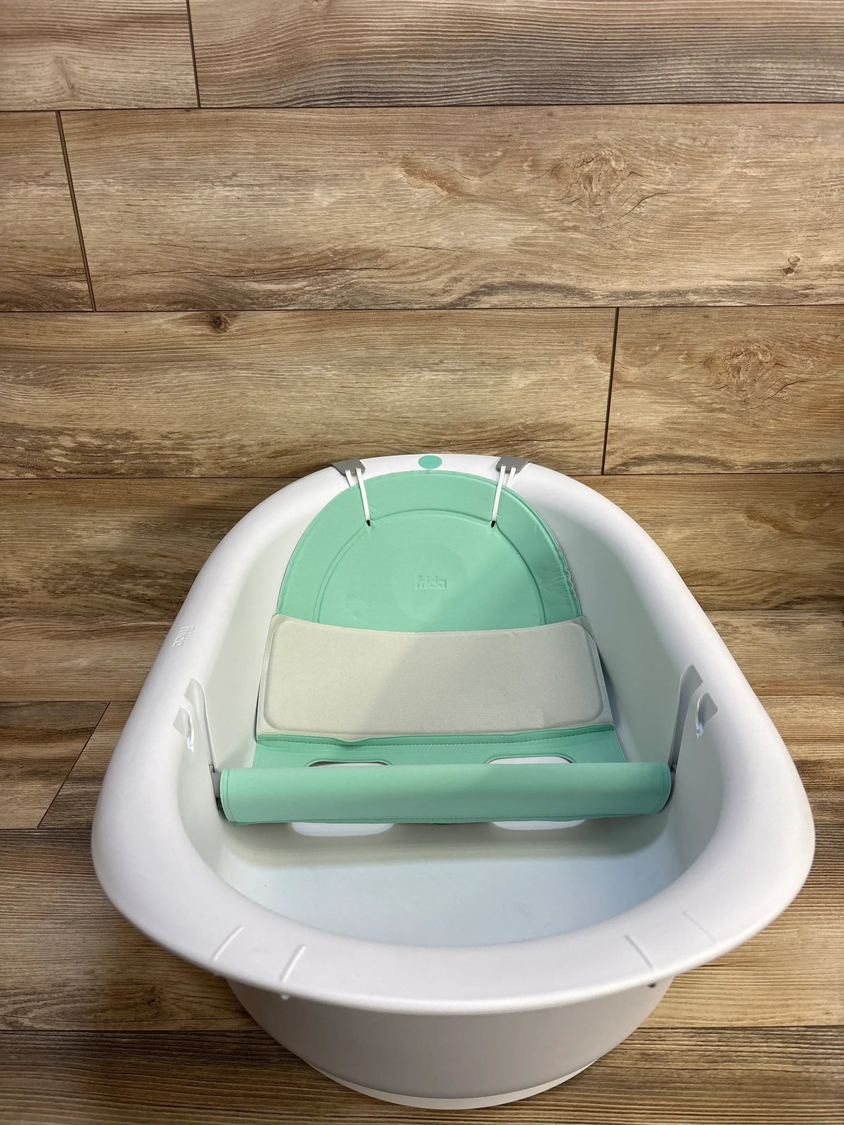 Front view of Frida Baby 4-in-1 Grow-With-Me Bath Tub in white with mint infant support and safety bar on wood floor