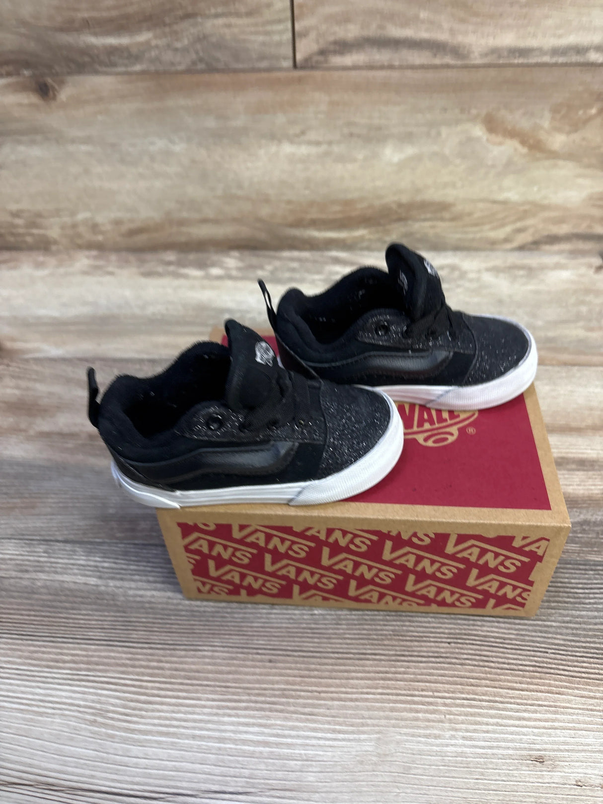 Angled view of black Vans Knu Skool sneakers showcasing glitter upper and white rubber soles on a Vans box on wood surface