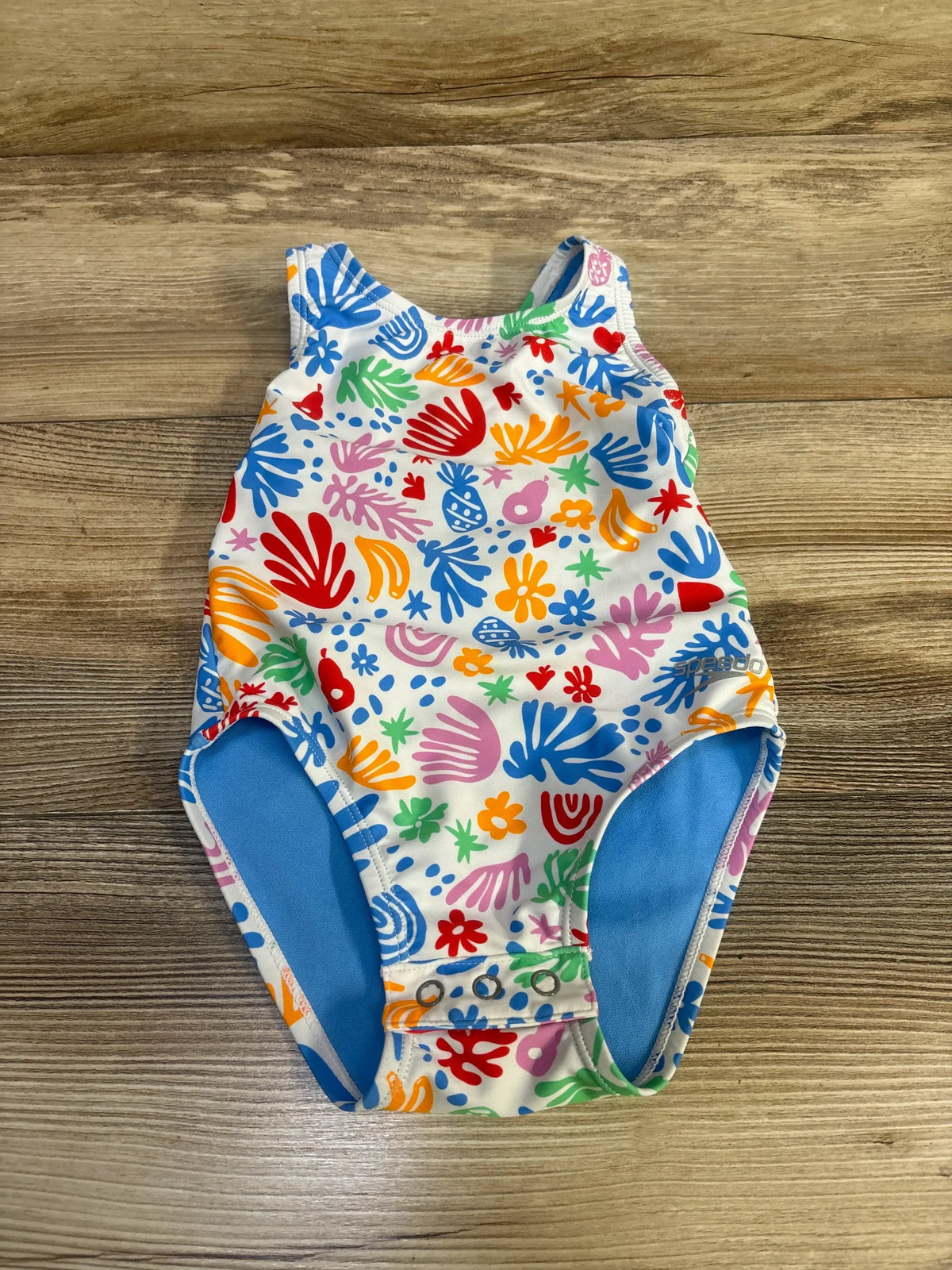 Front view of Speedo one-piece swimsuit size 2T, white with multicolored print and crotch snap closures on wood