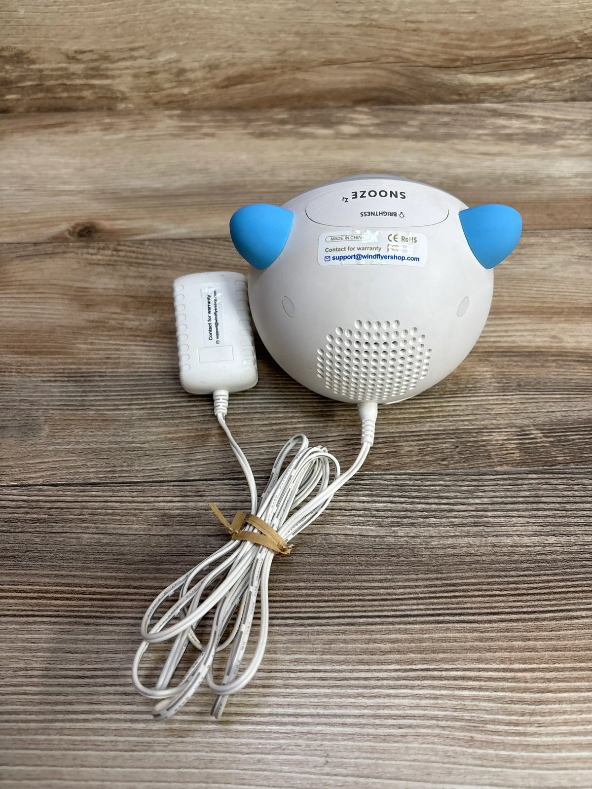 Back view of WindFlyer OK-to-wake clock showing blue ears, speaker grille, and power adapter on wood floor