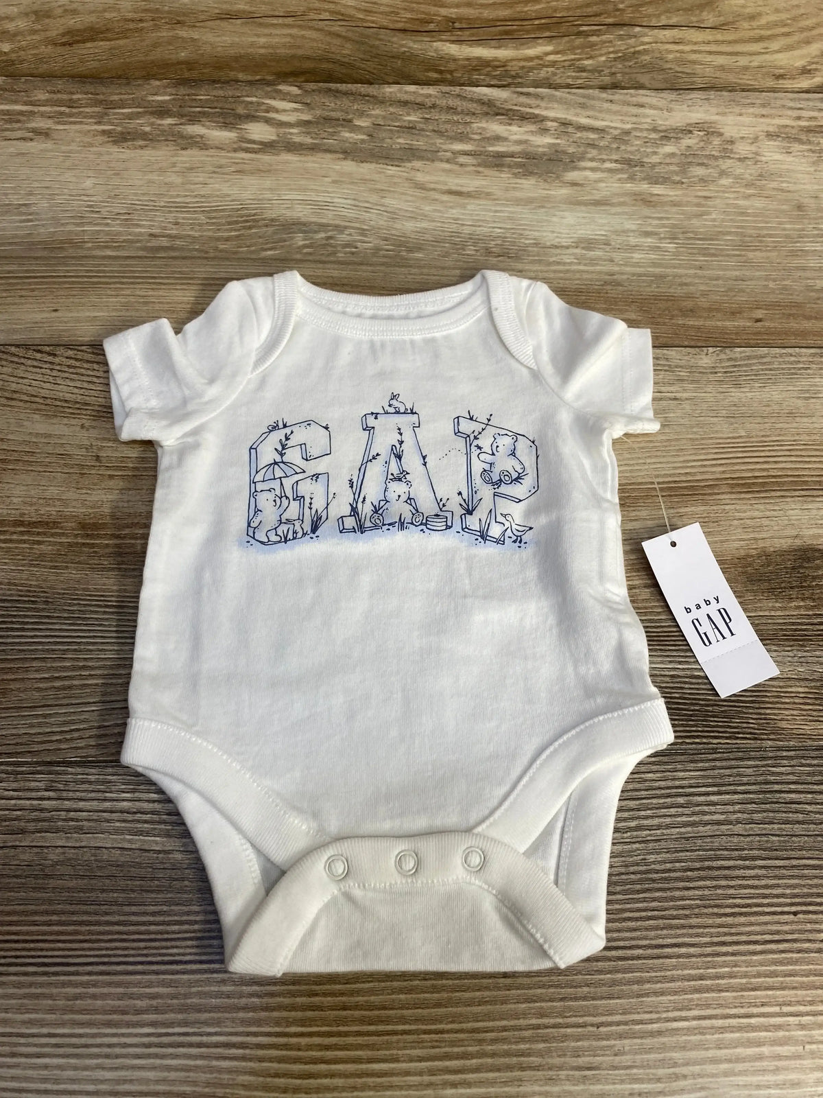 Front of white newborn Baby Gap bodysuit with blue GAP animal print letters, three snaps, and tag on wood surface