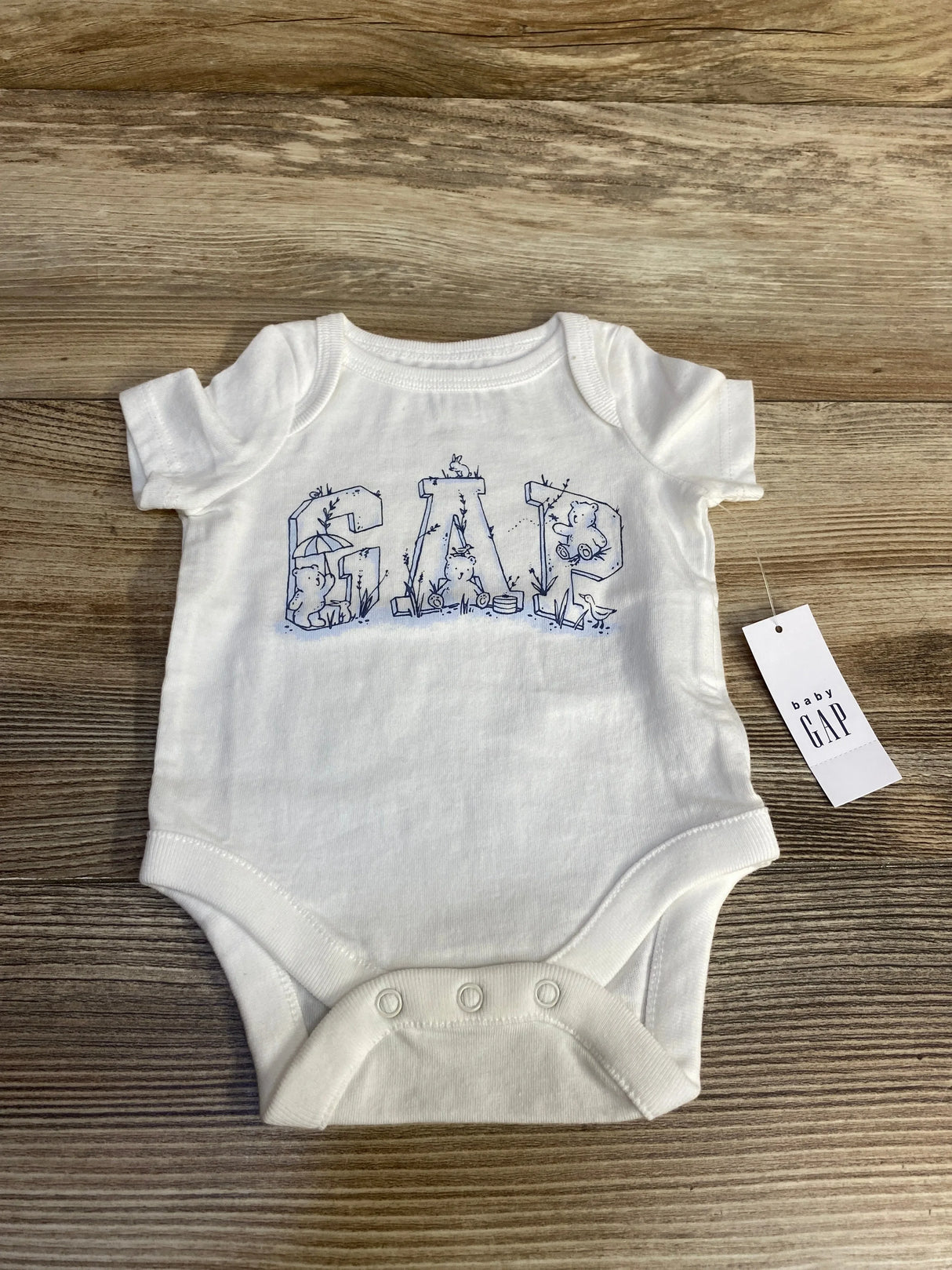 Front of white newborn Baby Gap bodysuit with blue GAP animal print letters, three snaps, and tag on wood surface