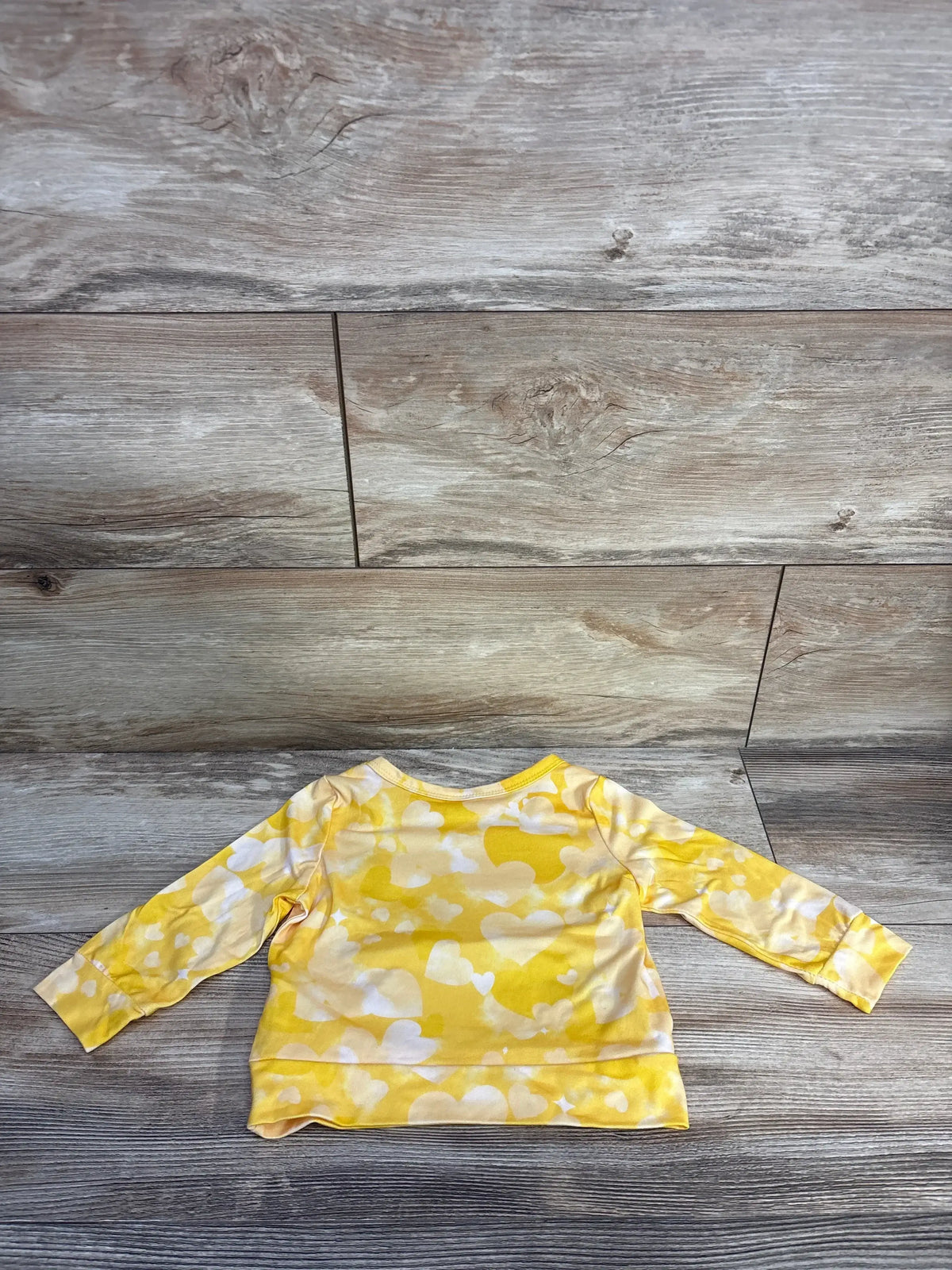 Back view of yellow baby long-sleeve shirt showcasing white heart print and banded hem on wood plank background
