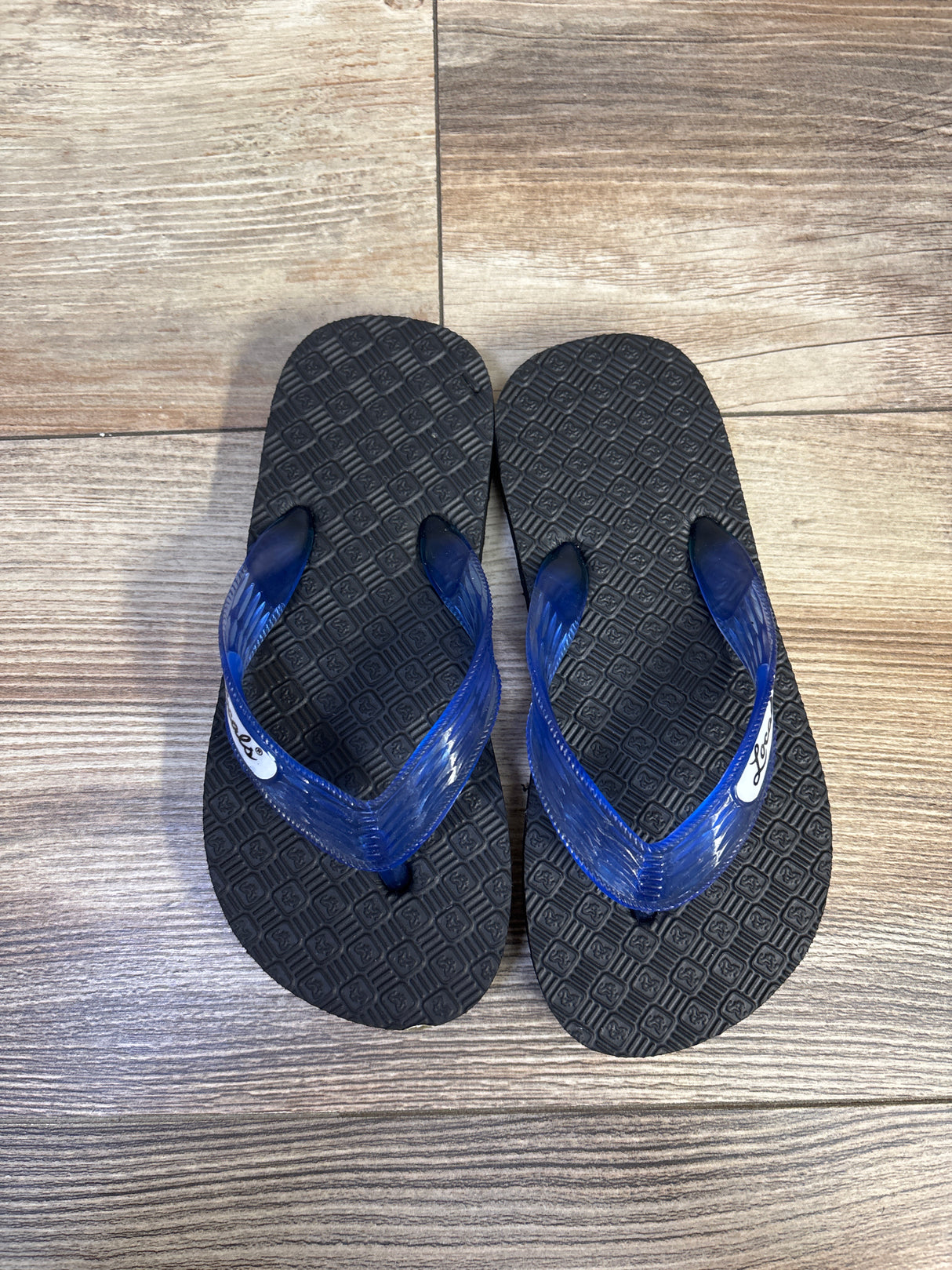 Top-down view of Locals Original flip flops size 7.5C showcasing translucent blue straps and textured black footbeds on wood floor