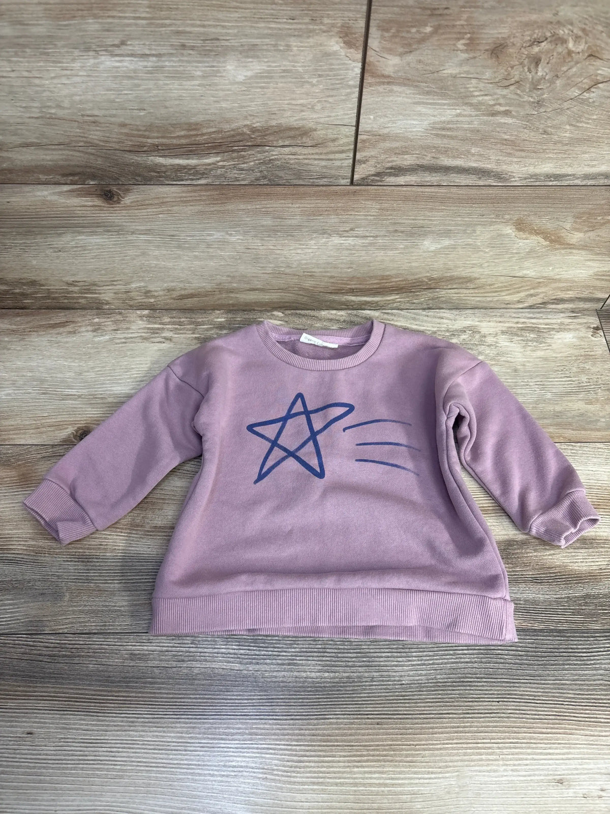 Flat lay of mauve oversized organic toddler sweatshirt showing blue shooting star graphic, ribbed cuffs and hem on wood floor