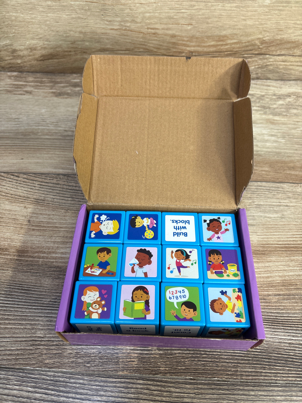 Top-down view of Lakeshore Calm Down Cubes, 12 blue illustrated emotion cubes in an open purple box on wood