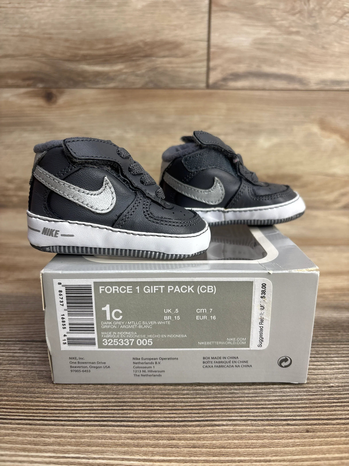 Dark grey infant Nike Force 1 shoes with silver swoosh on original box, wooden background.
