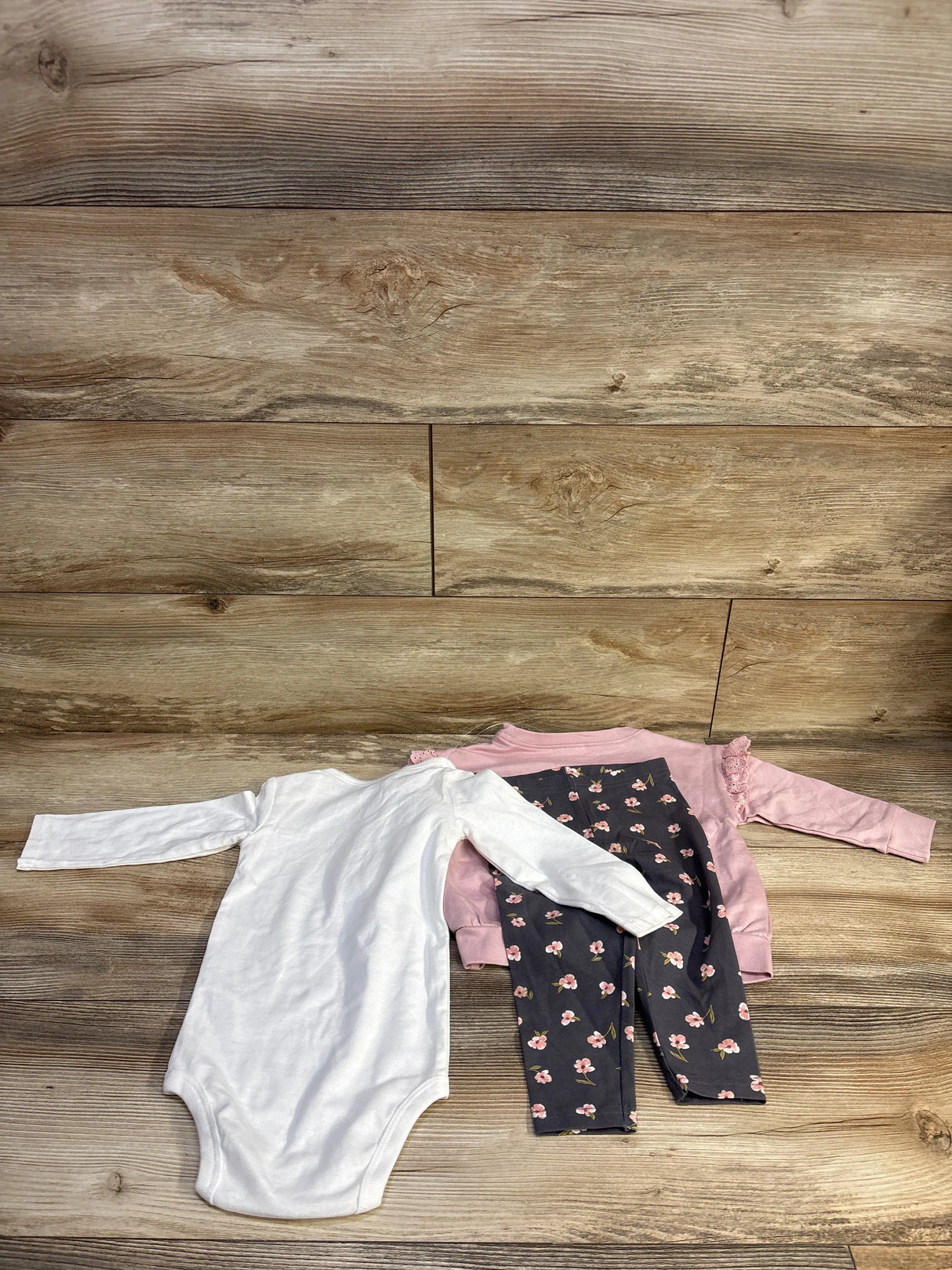 Flat lay of 3-piece baby set with white long-sleeve bodysuit, pink ruffle shoulder top, and navy floral leggings on wood background