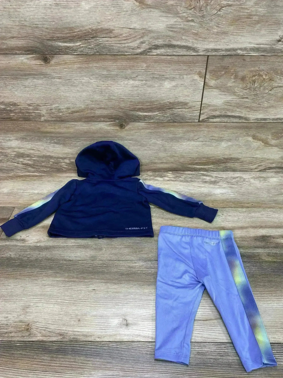 Flat lay of Nike 2-piece Therma-FIT full-zip navy hoodie and lavender leggings size 6M showing hood, ombre side stripes, and elastic waistband on wood floor