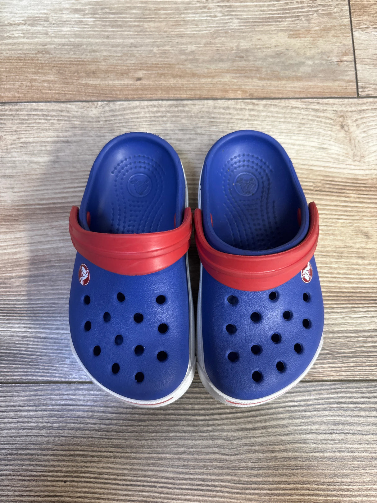 Top view of Crocs Crocband II clogs showing blue uppers, red heel straps, white midsoles and ventilation holes, child size 6/7 on wood floor