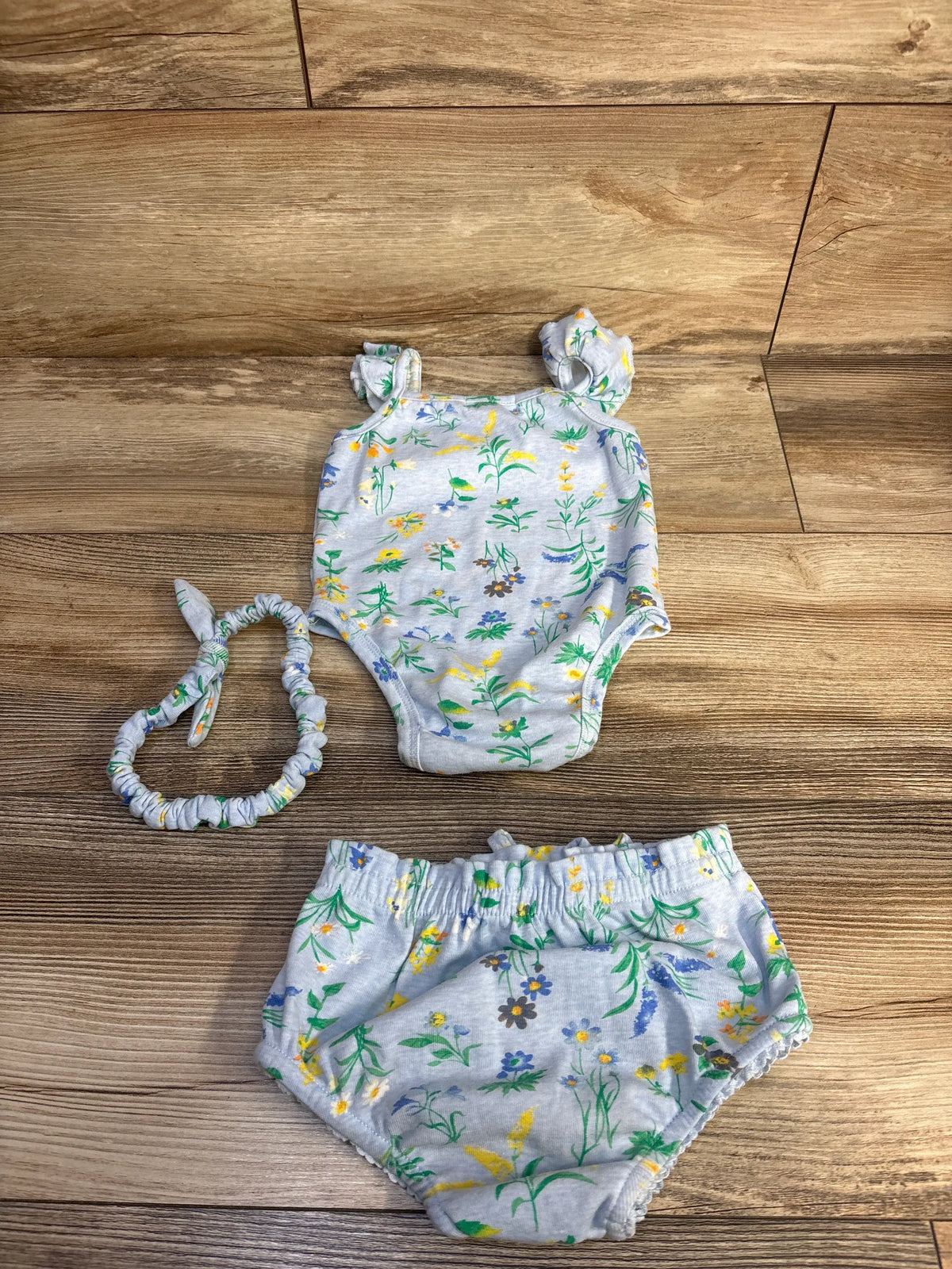 Top-down view of Btween baby 3pc bodysuit, bloomers, and headband set showcasing a blue floral print on wood flooring