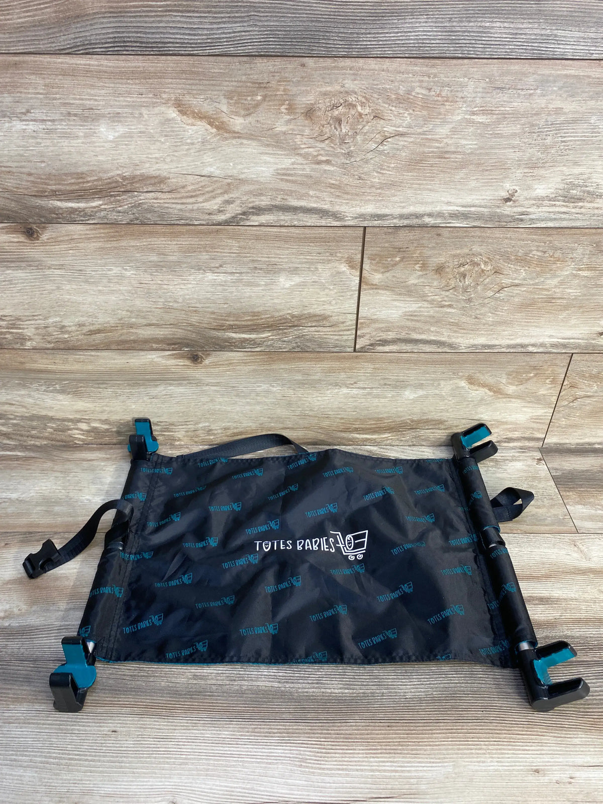 Top-down view of Totes Babies shopping cart hammock: black fabric with TOTES BABIES logo, adjustable straps and clips on wood