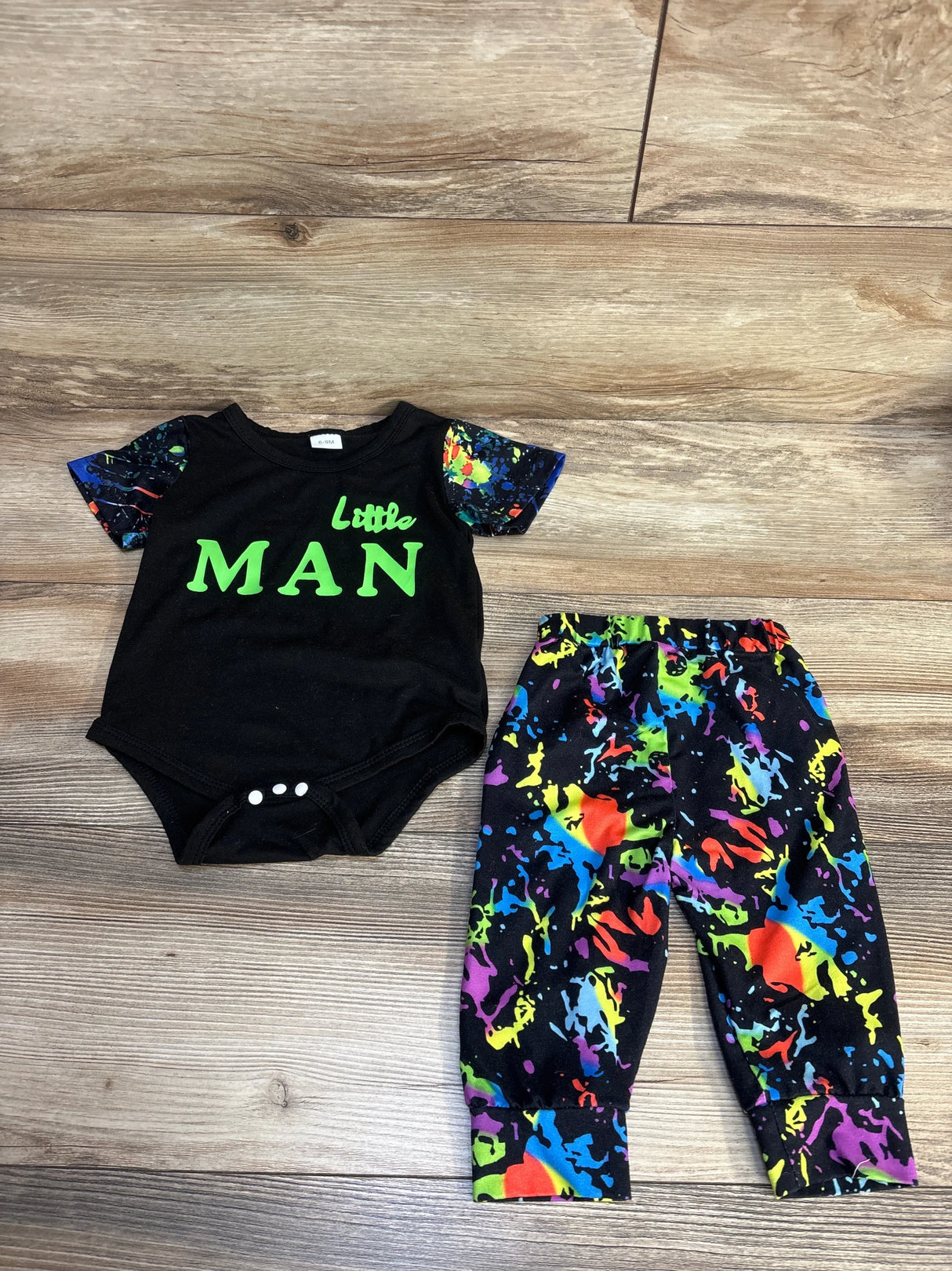 Flat lay of black baby bodysuit with neon green Little MAN text and matching multicolor paint-splatter pants on wood floor