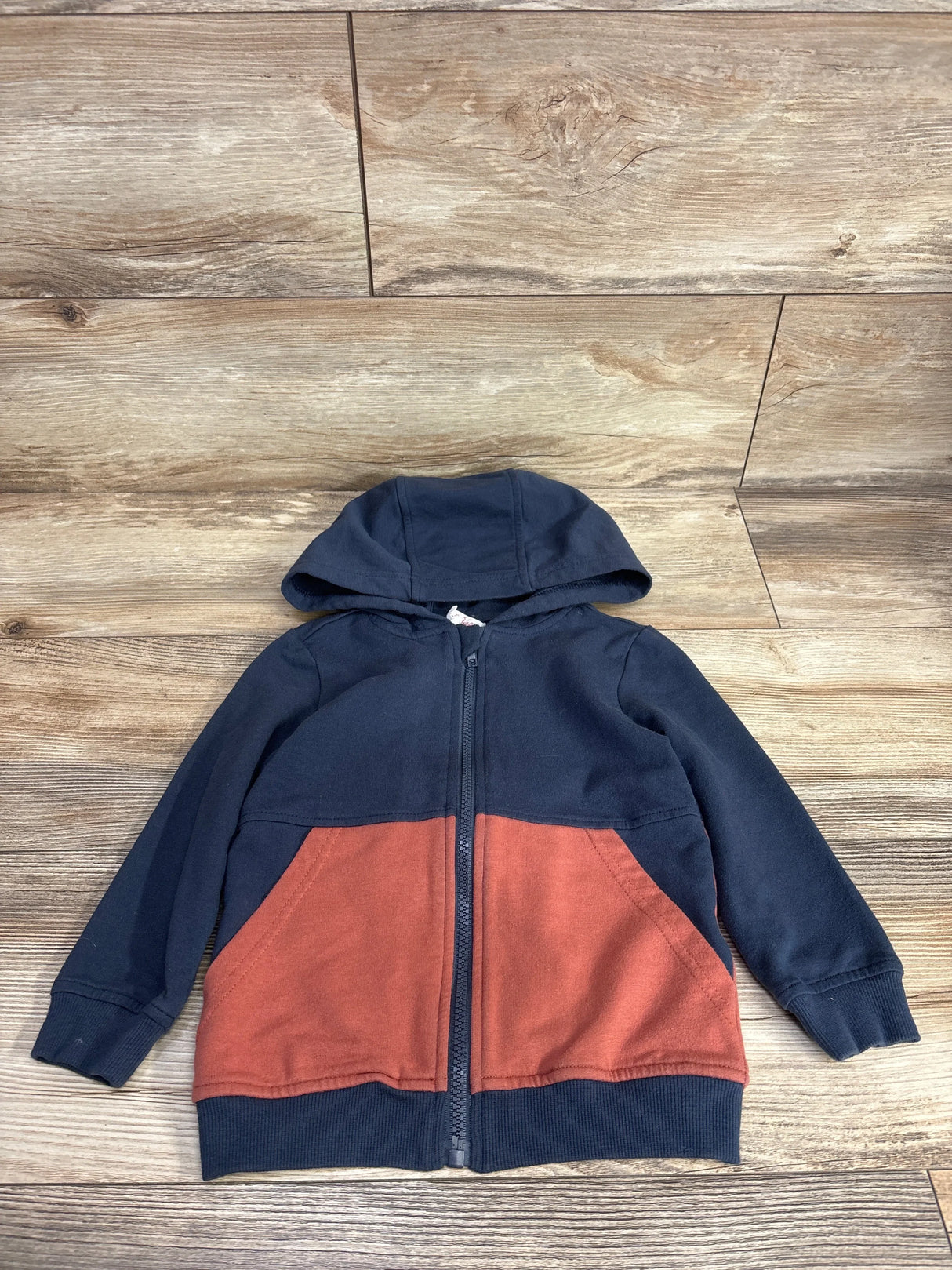 Kids zip-up hoodie with navy blue top, rust orange pockets and hood, laid on wood floor