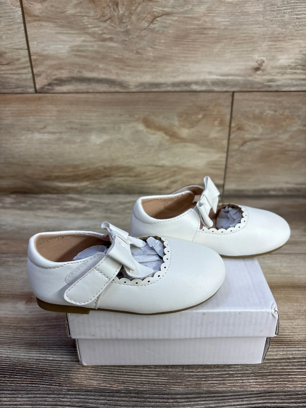 Side view of white leather baby Mary Jane flats size 7C showcasing scalloped trim and bow on white box, wood floor background