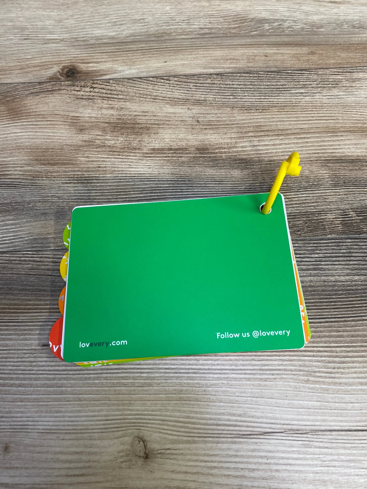 Top-down view of Lovevery The Thinker Play Guide cards with green back, multicolored tabbed edges and yellow key ring on wood surface