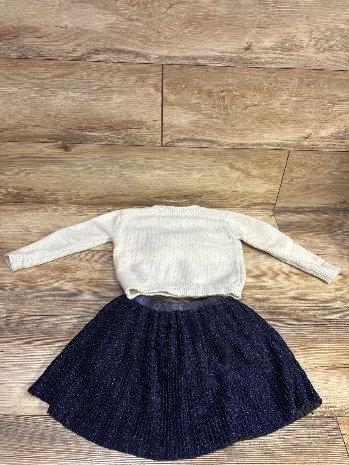 Flat lay of Lands’ End toddler knit white sweater and navy pleated skirt on wood floor