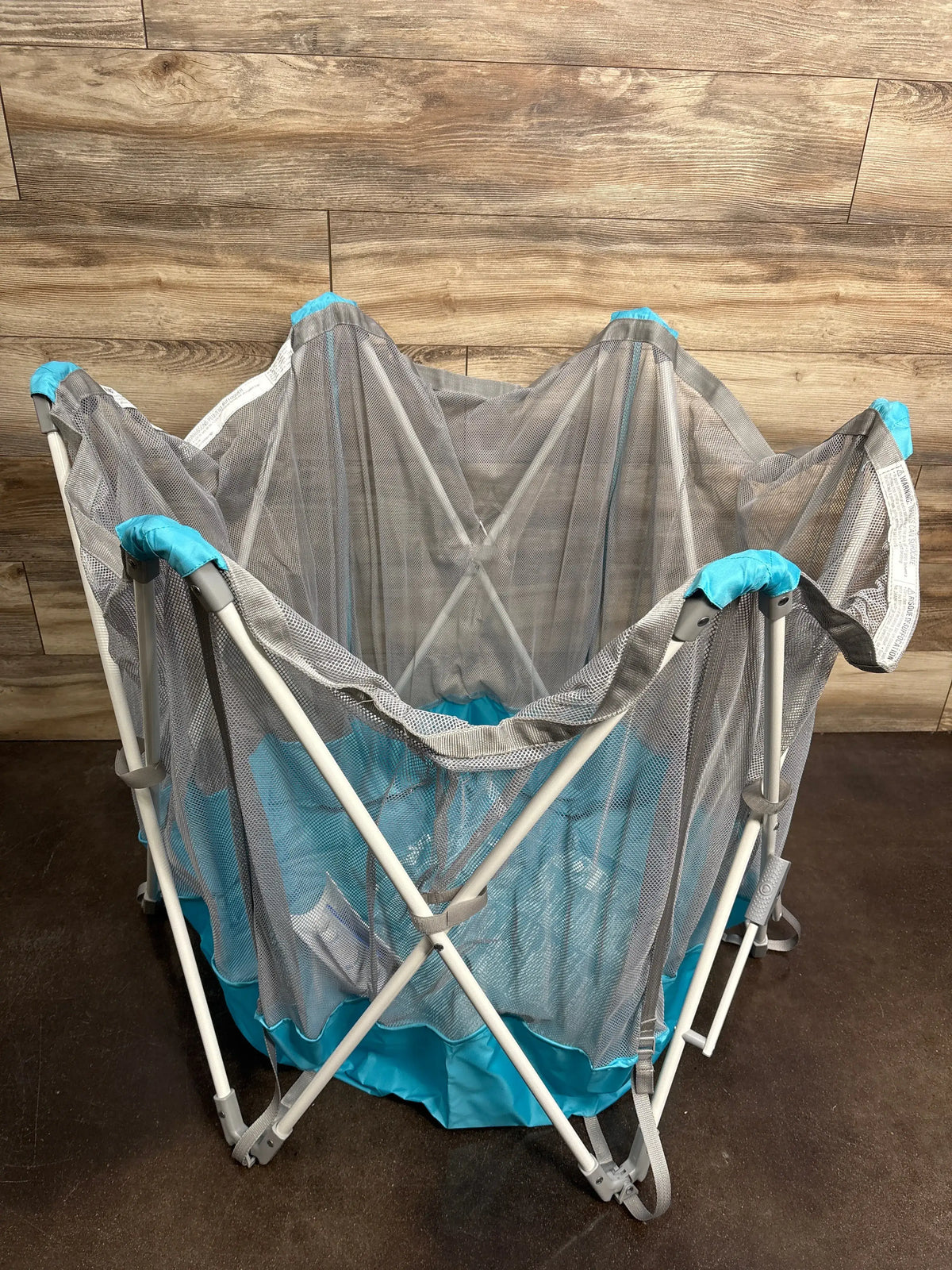 Front view of Regalo My Play portable 6-panel play yard showcasing teal mesh walls and nylon bottom with a wood-patterned background