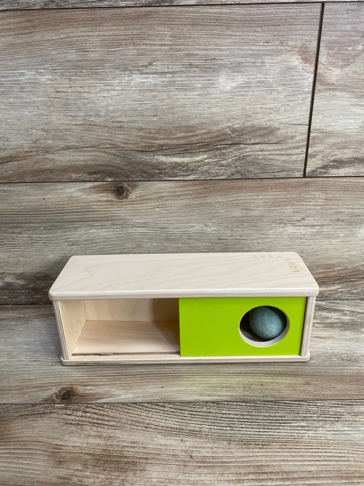 Front view of Lovevery Sliding Top Box showing lime green sliding panel with circular hole and blue felt ball against wood background
