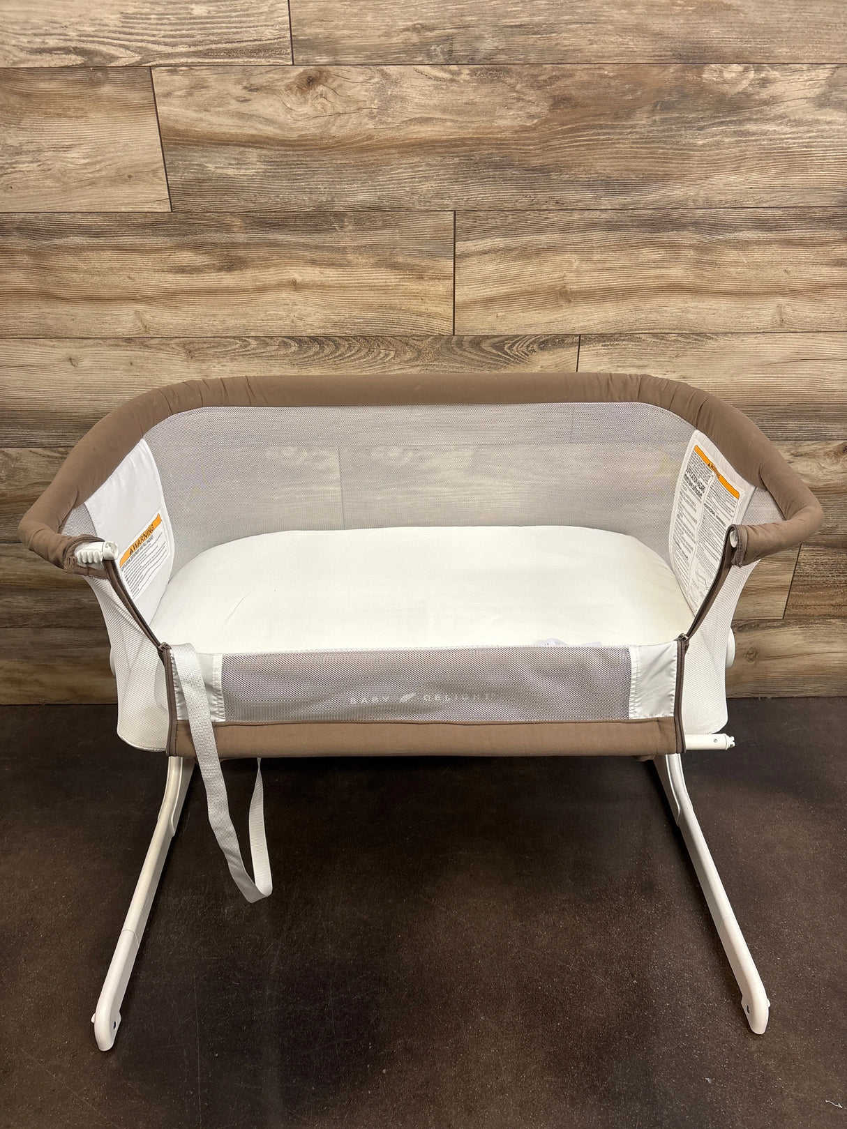 Front view of Baby Delight Beside Me Dreamer bassinet in mocha with mesh sides and mattress on wood backdrop