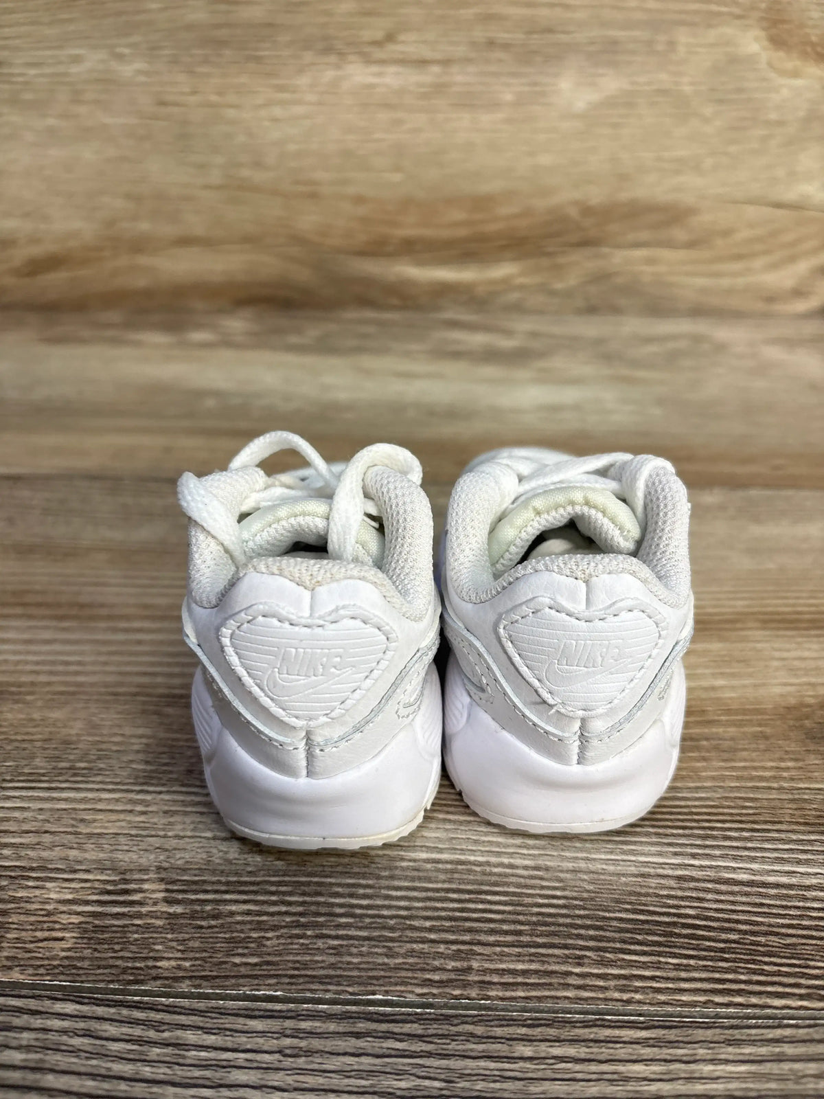 Rear view of Nike Air Max 90 TD Triple White sneakers, size 2C, showing Nike heel logos on a wood floor