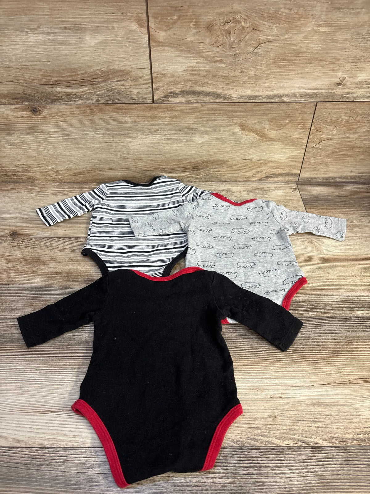 Top view of Baby Guess 3pk bodysuits, size 0 to 3 months, with long sleeves, varied patterns, and red trim on wood floor